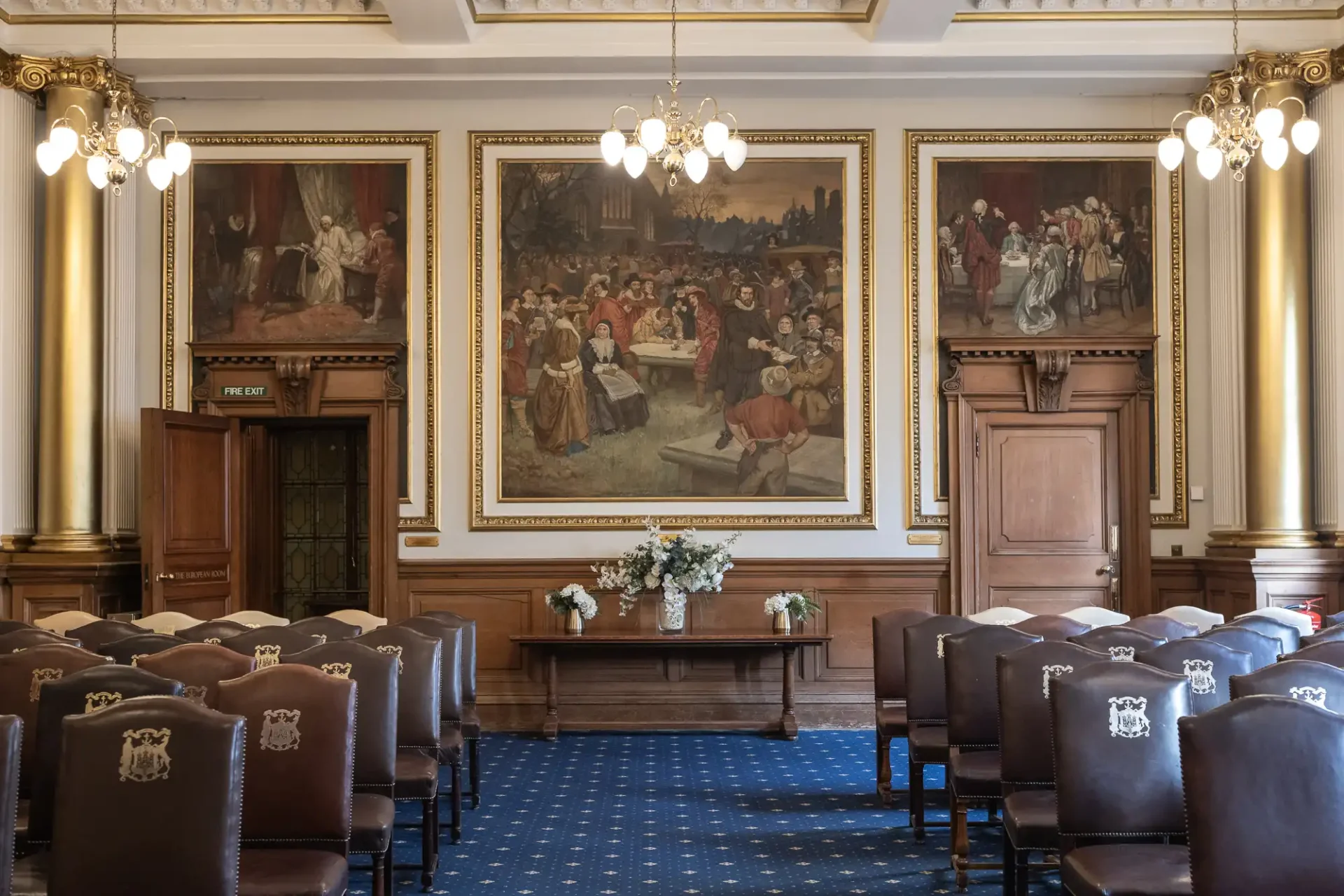 Ornate room with paintings, chandeliers, and rows of leather chairs facing a table with floral arrangements.