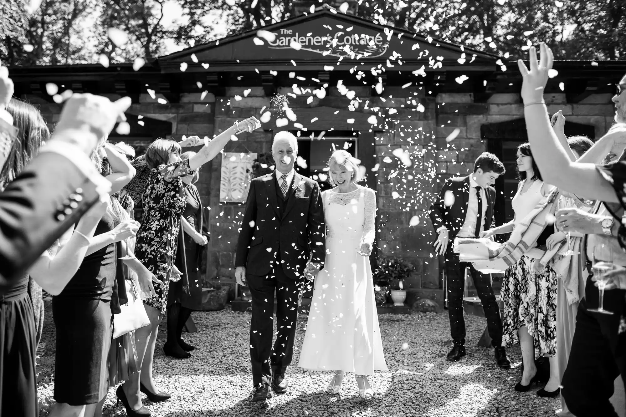 A bride and groom walk down a gravel path while guests throw confetti around them outside The Gardener's Cottage.