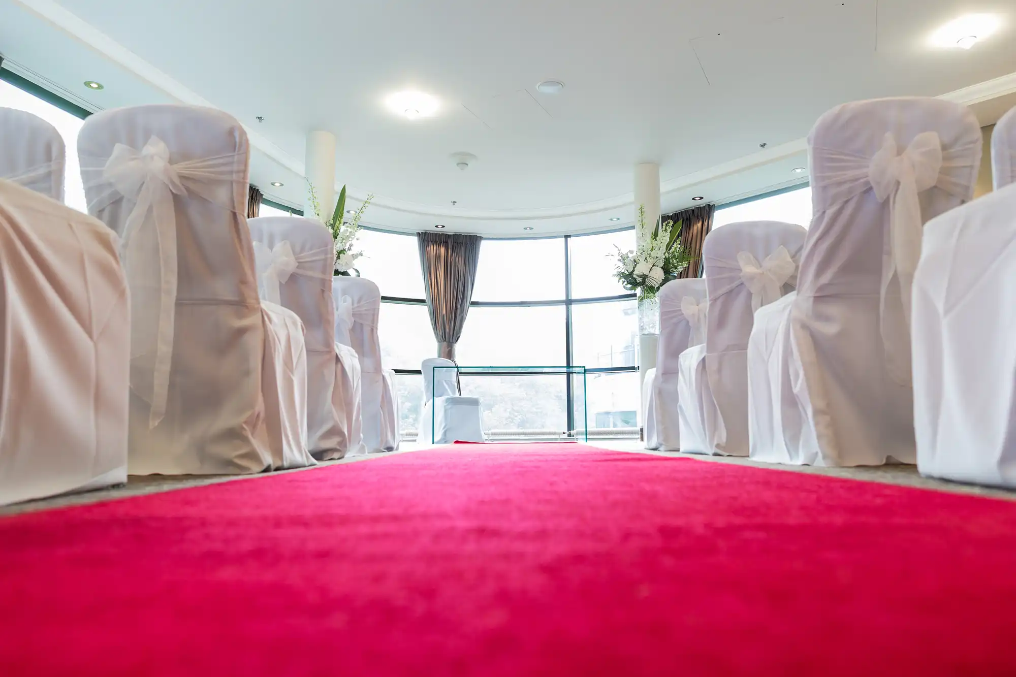 Low-angle view of a red carpet aisle in a wedding venue with rows of white chair covers and decorative bows.