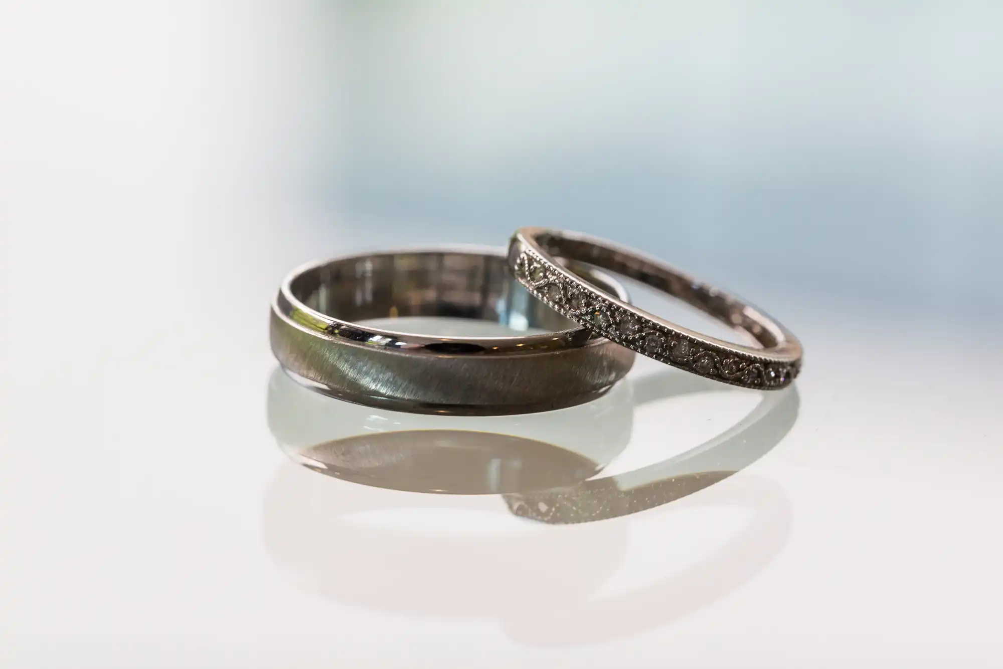 Two wedding rings, one encrusted with tiny diamonds and the other a smooth, dark metal, on a reflective surface with a blurred white background.