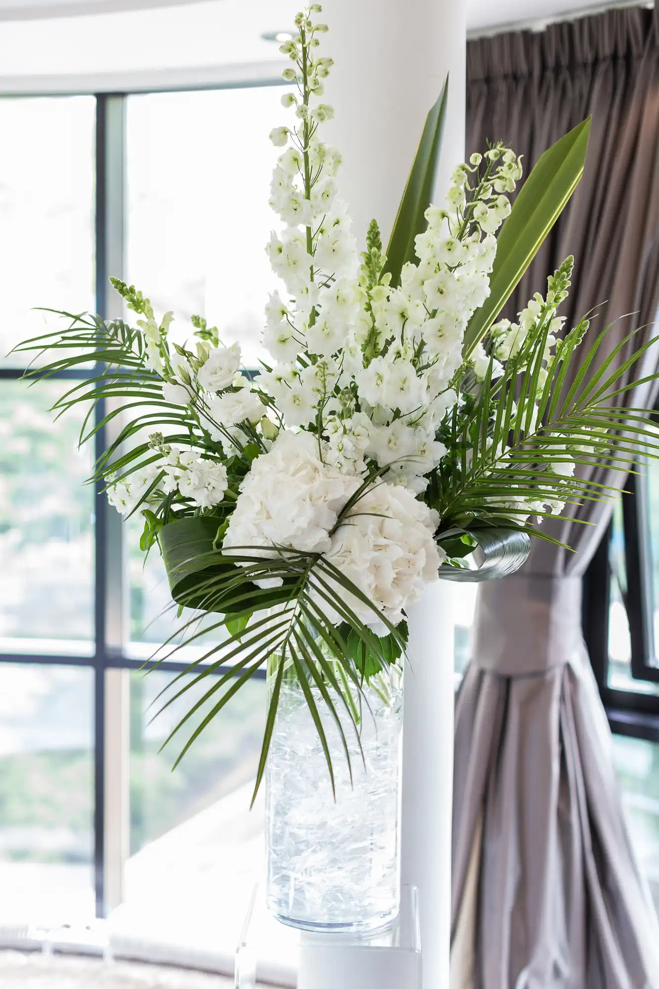 Elegant floral arrangement featuring white hydrangeas, tall snapdragons, and tropical palm leaves in a clear vase, set against a window with curtains.
