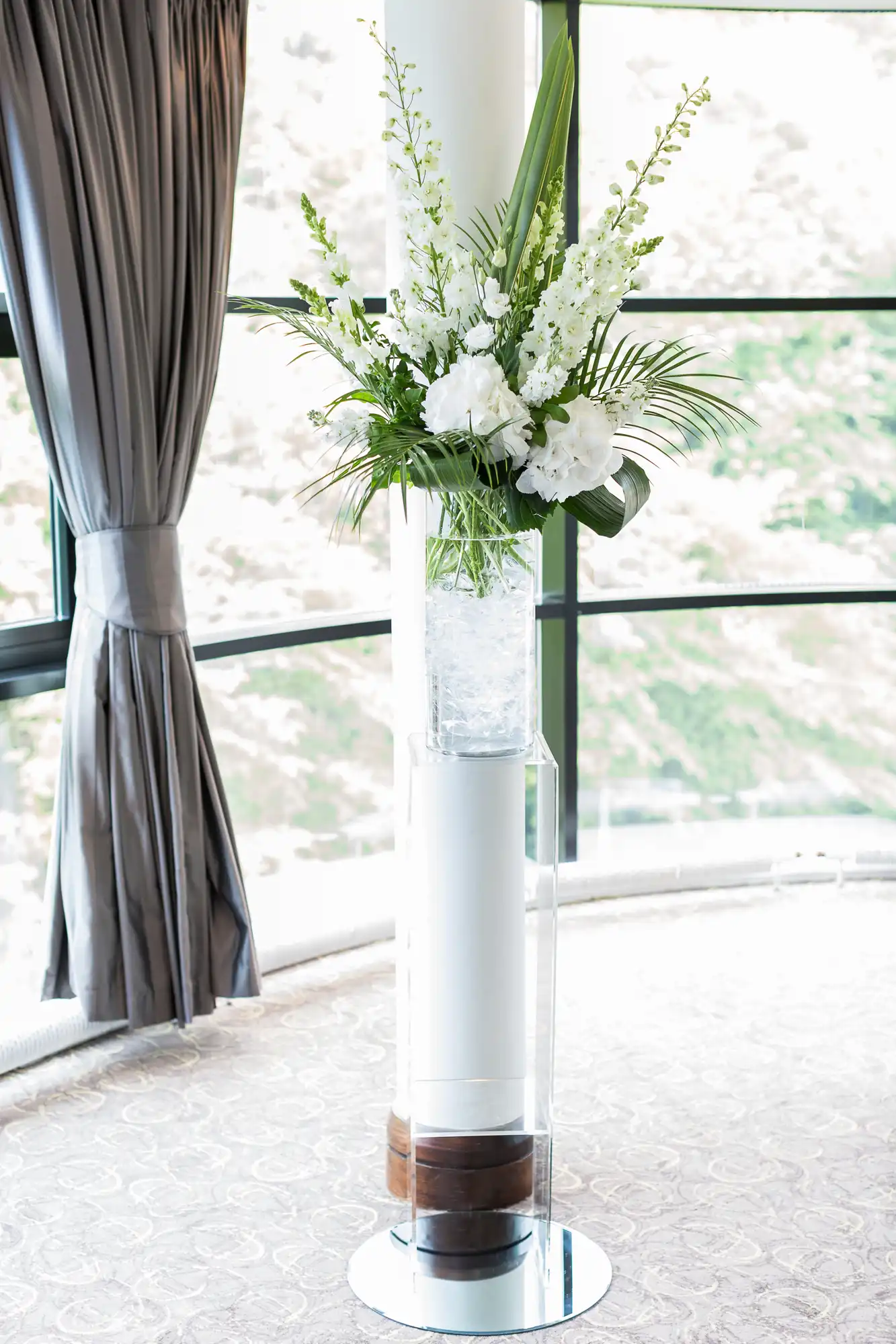 Elegant floral arrangement with white flowers and green leaves in a tall, clear vase on a pedestal near a curtained window.