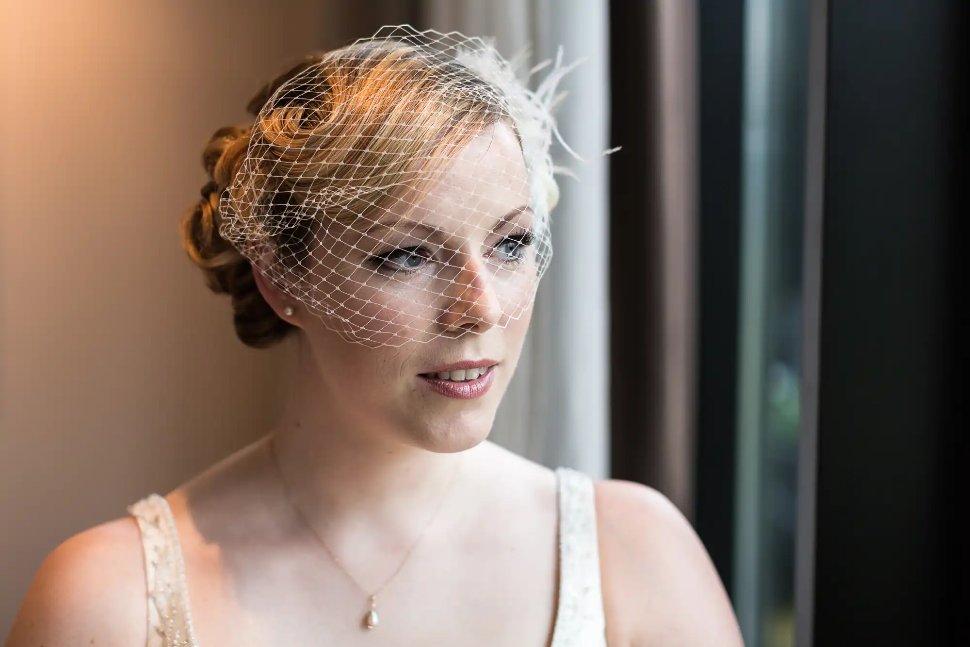 A bride wearing a vintage-inspired birdcage veil looks contemplatively out a window, her hair in an elegant updo.