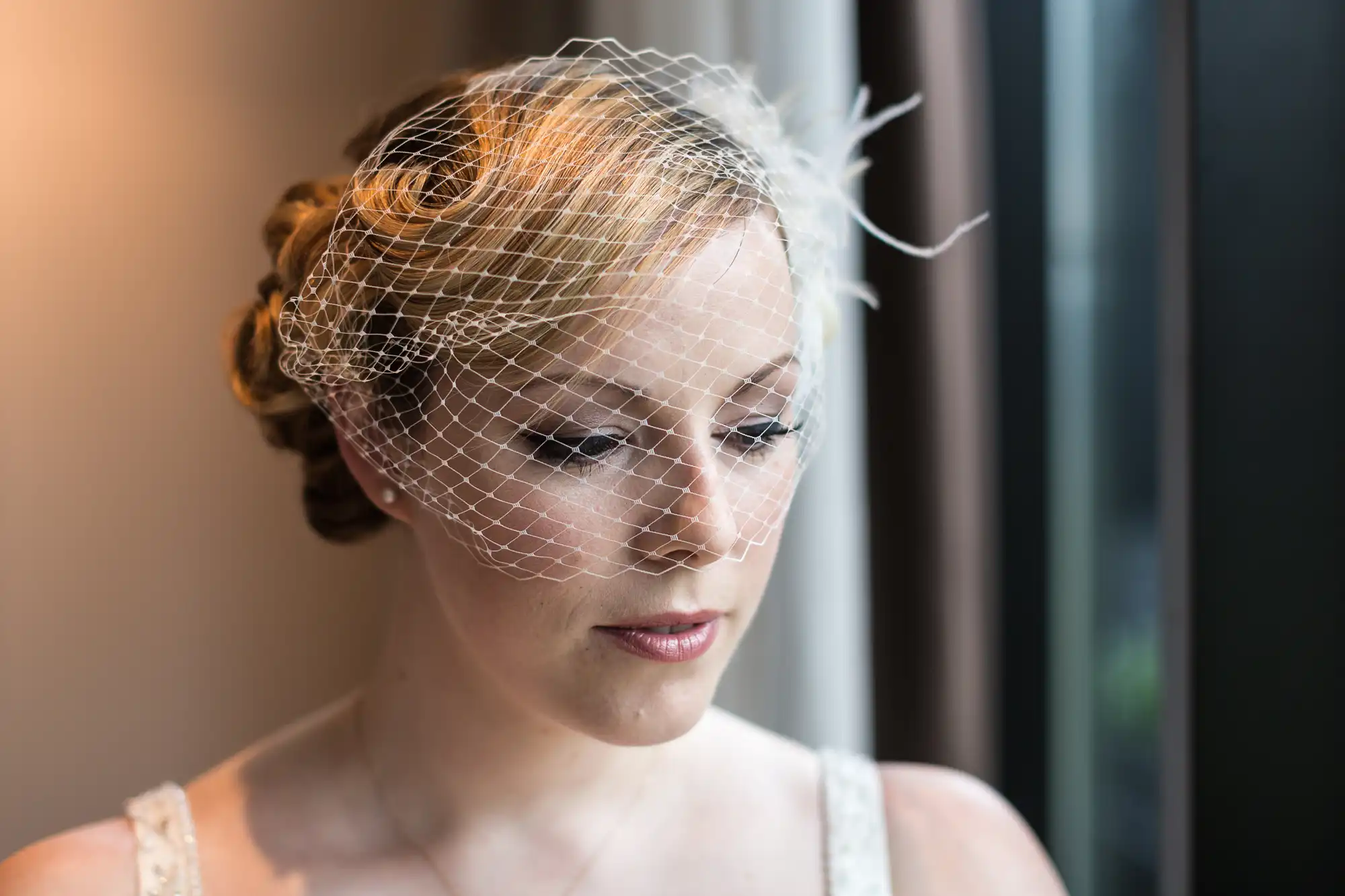 A bride in a white dress and birdcage veil gazes out a window, natural light illuminating her thoughtful expression.
