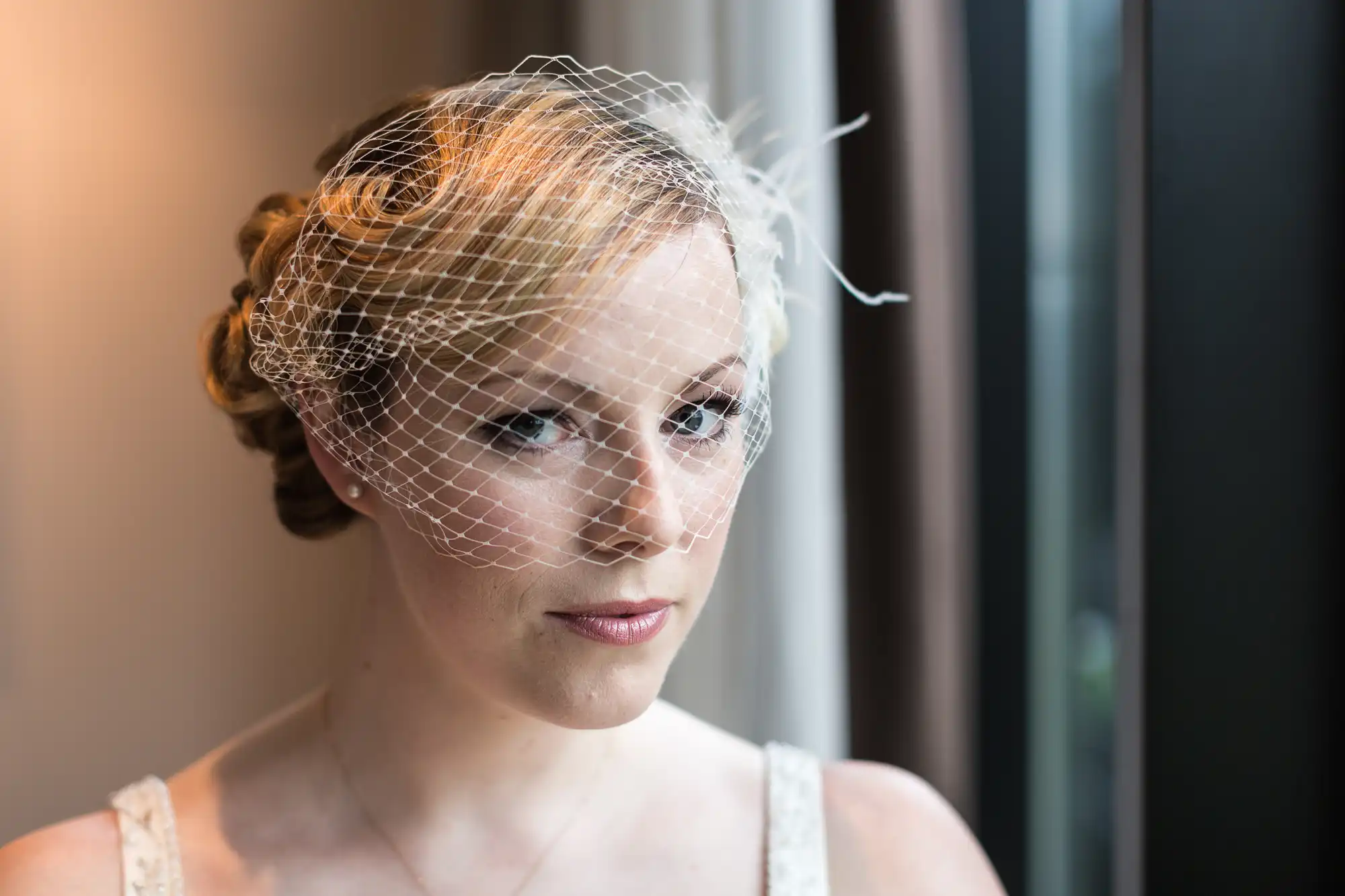 A bride wearing a veil gazes out a window, her elegant hairstyle and thoughtful expression captured in soft lighting.