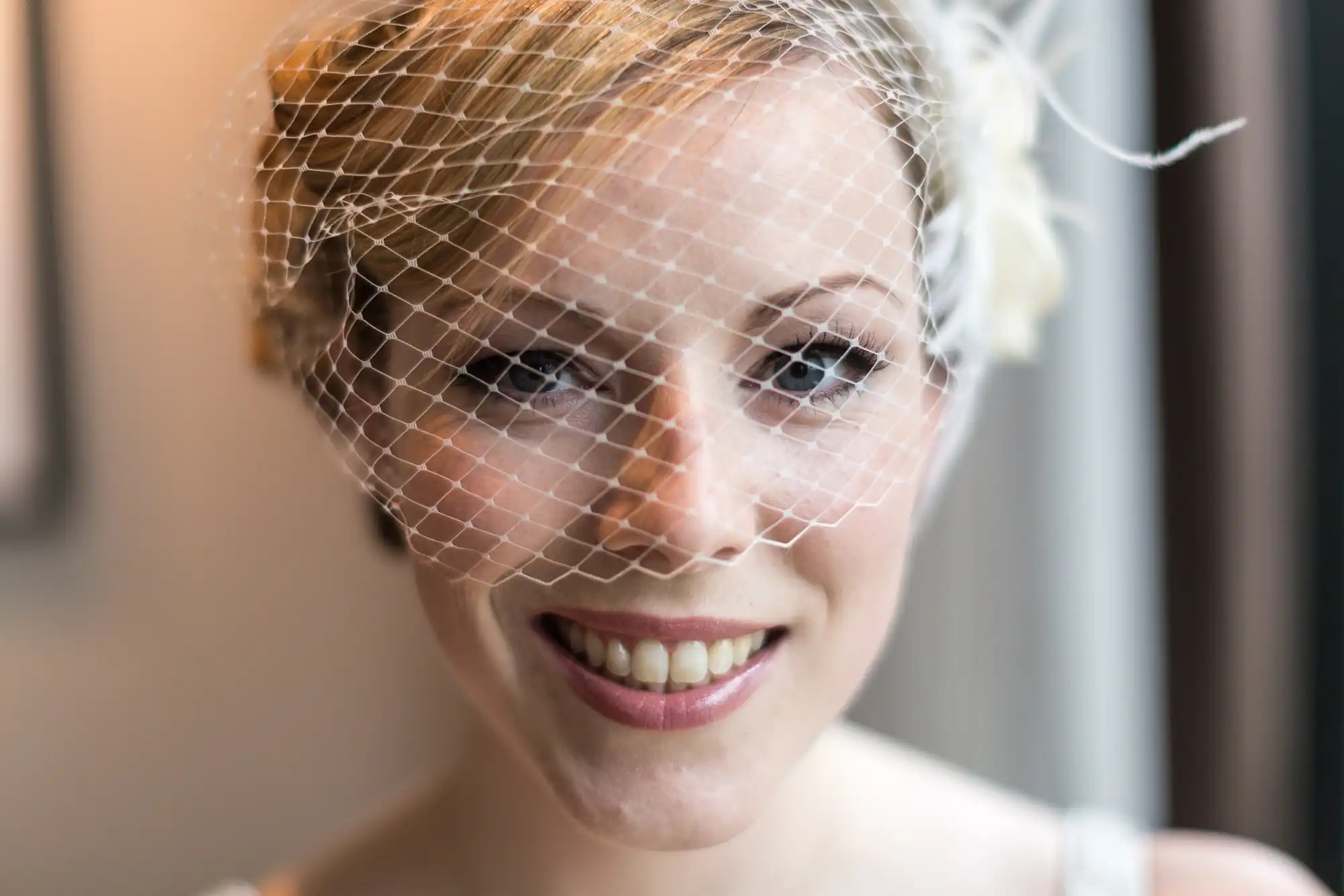 Close-up of a smiling woman with short hair wearing a white bridal veil with a mesh design, looking gently towards the camera.