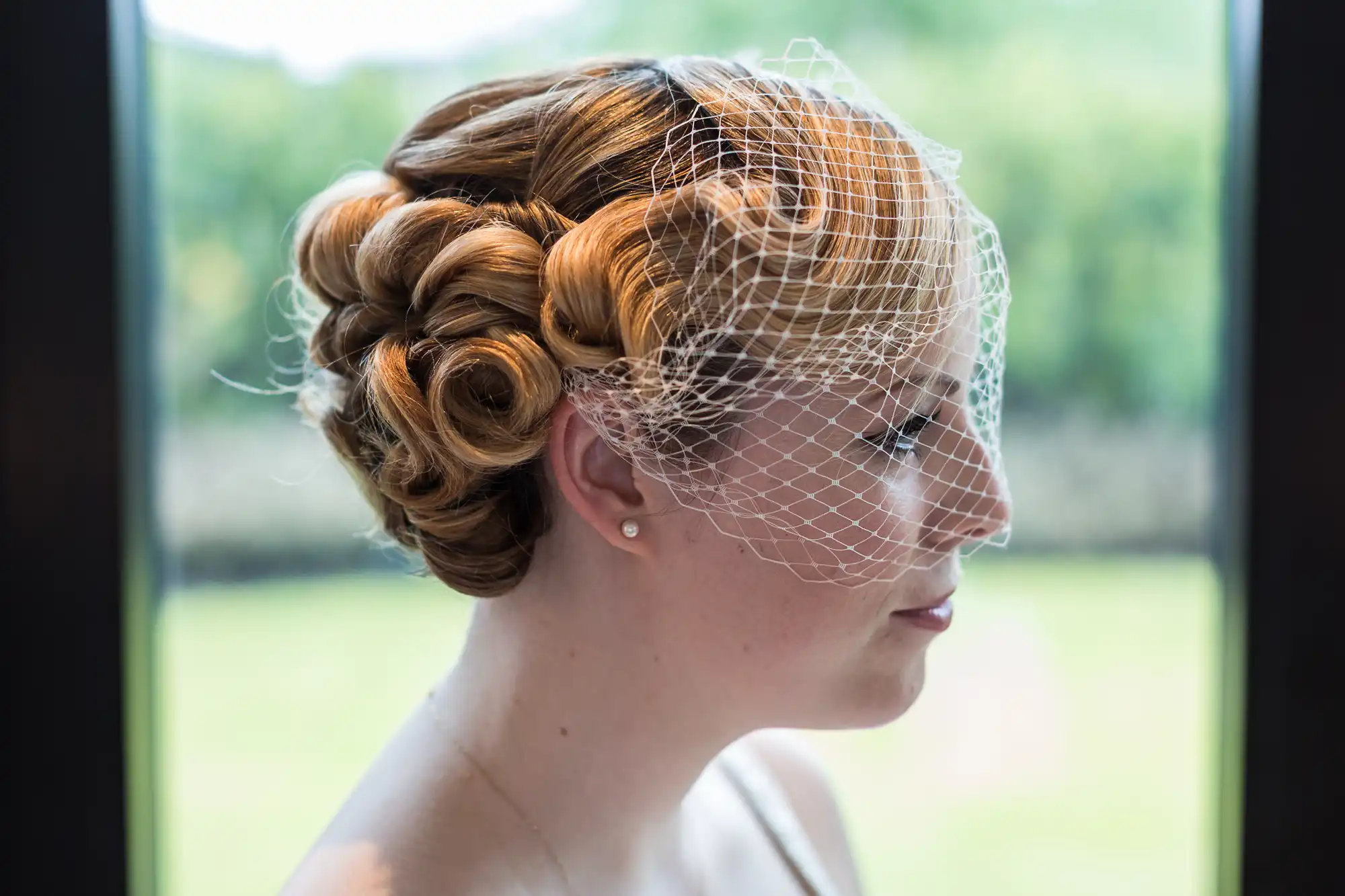 Profile of a woman gazing out a window, featuring an intricate updo hairstyle with a light birdcage veil.
