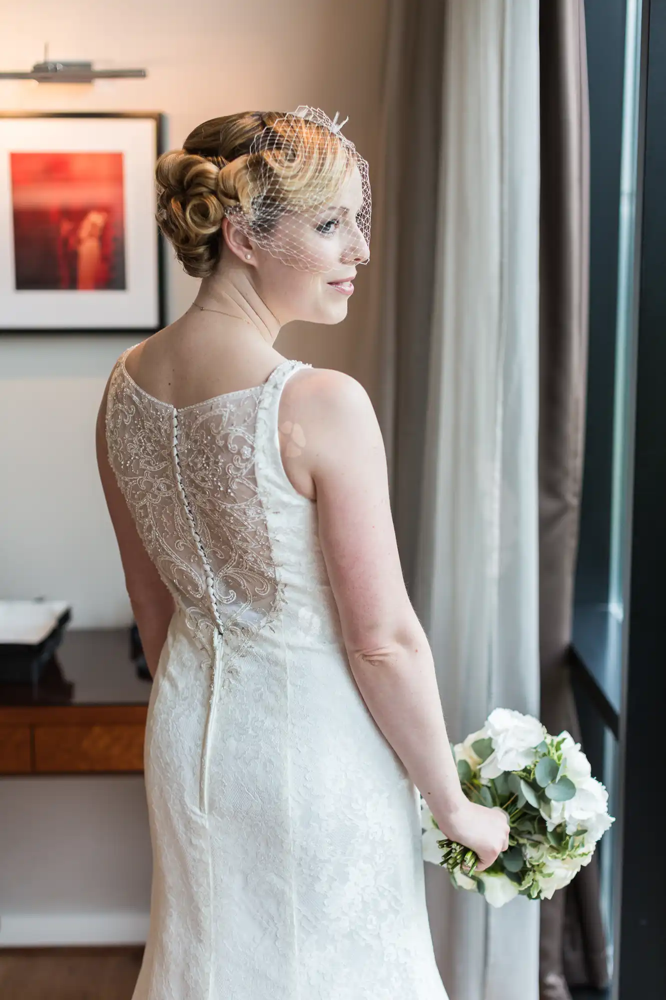 A bride in an elegant lace wedding dress looking out a window, holding a bouquet of white flowers, with a gentle smile.