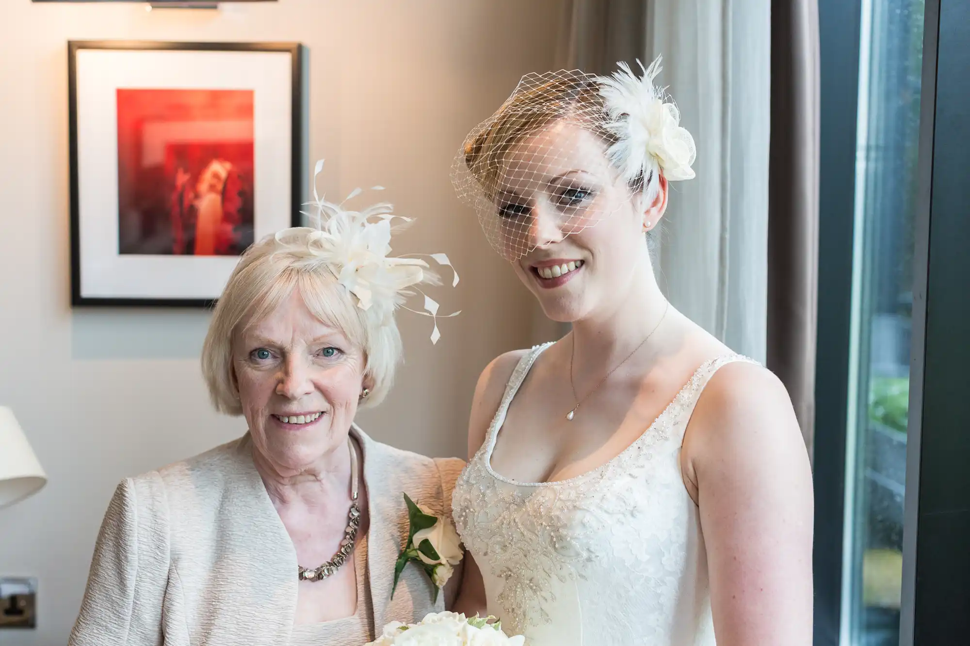A bride in a white dress and veil stands next to an older woman in a beige outfit, both smiling indoors.