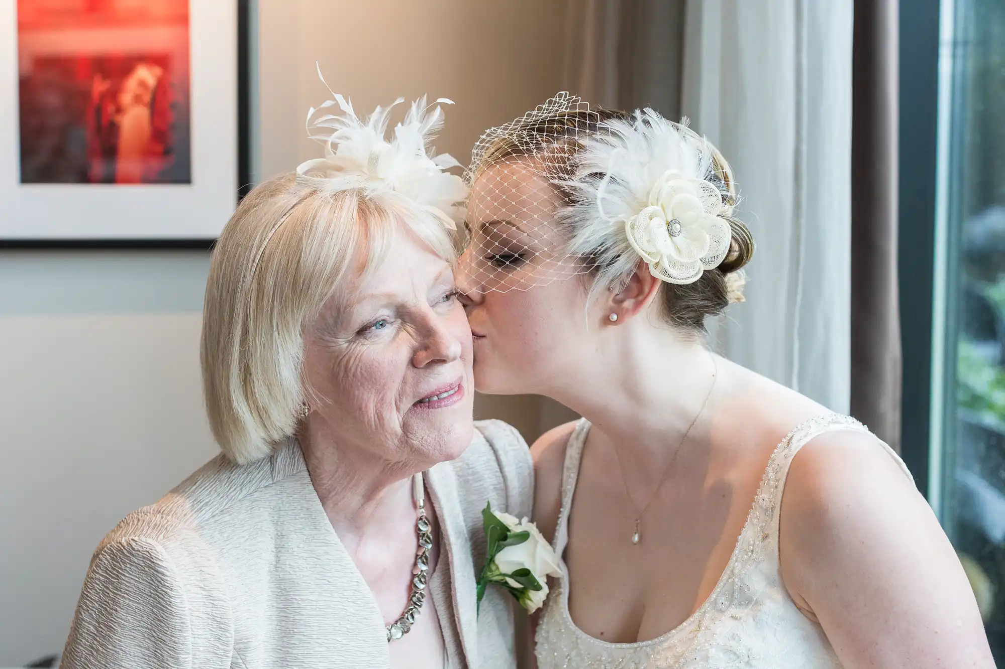 A bride in a white dress and hair accessory kisses an elderly woman on the cheek, both smiling happily indoors near a window.