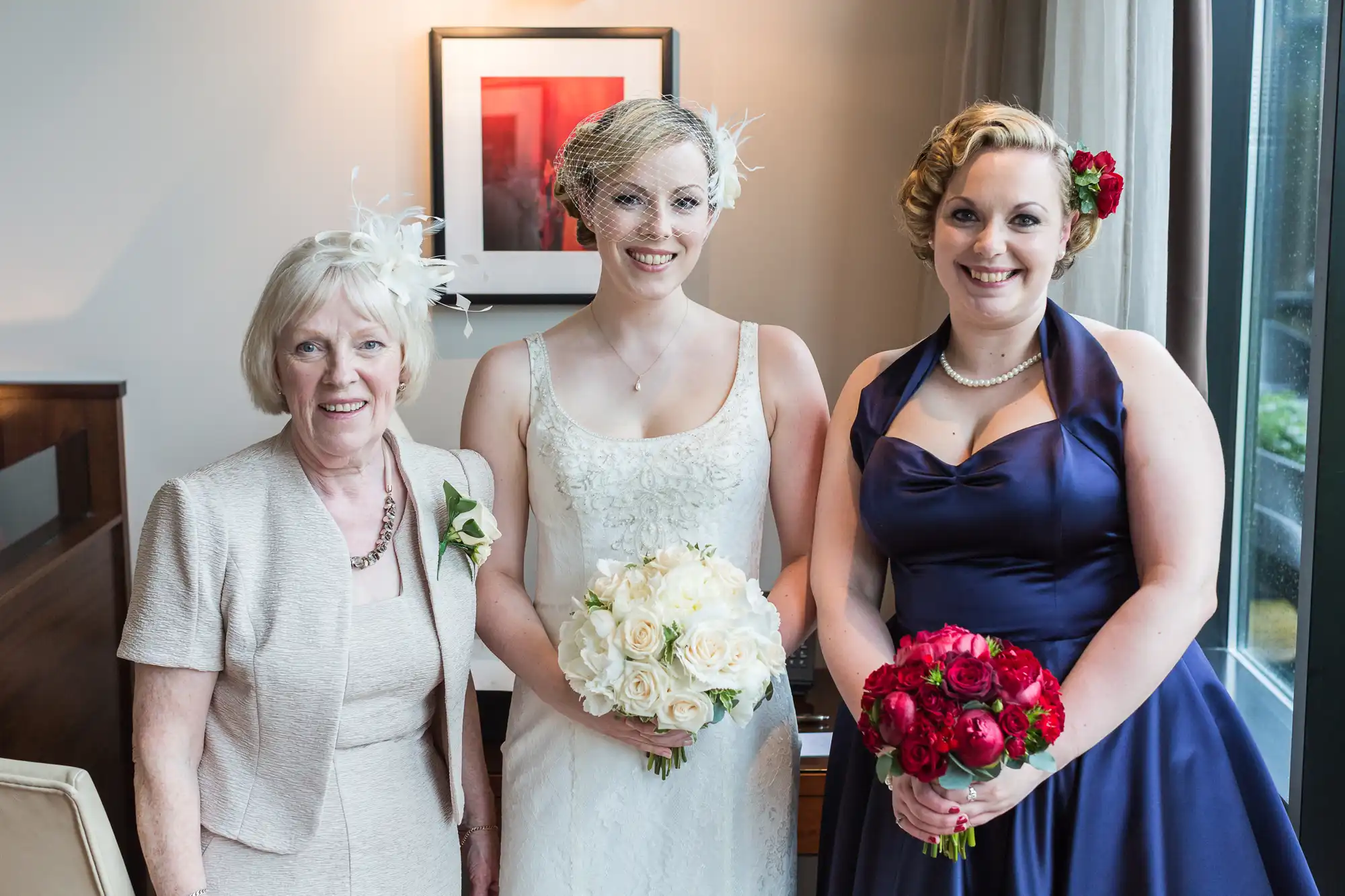 Three women in formal attire posing indoors, one in a wedding dress with a bouquet, flanked by two others in dresses, one holding red flowers.