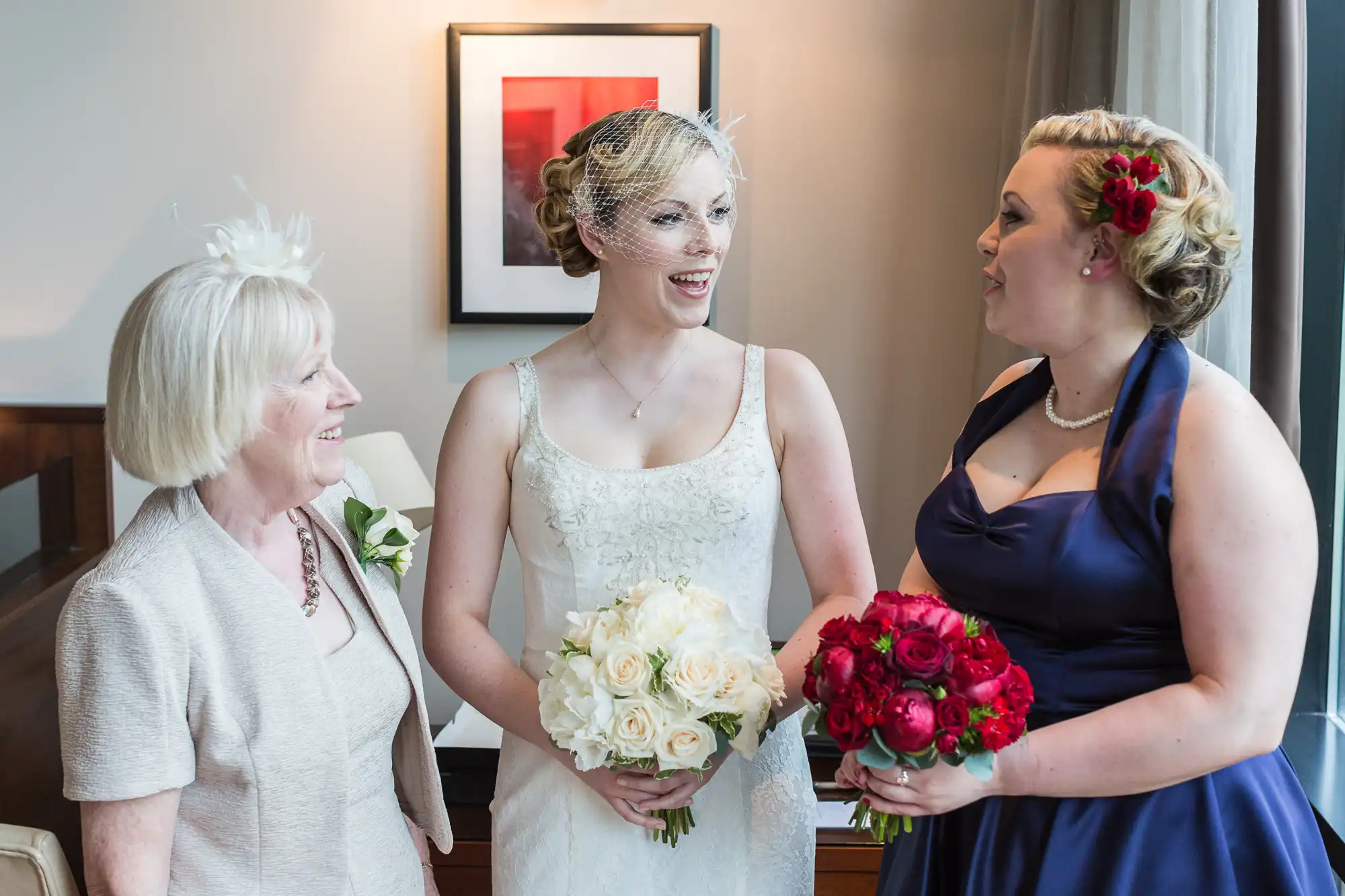 Three women at a wedding smiling and talking, with the bride in the center holding a bouquet of white roses and the other two holding red roses.
