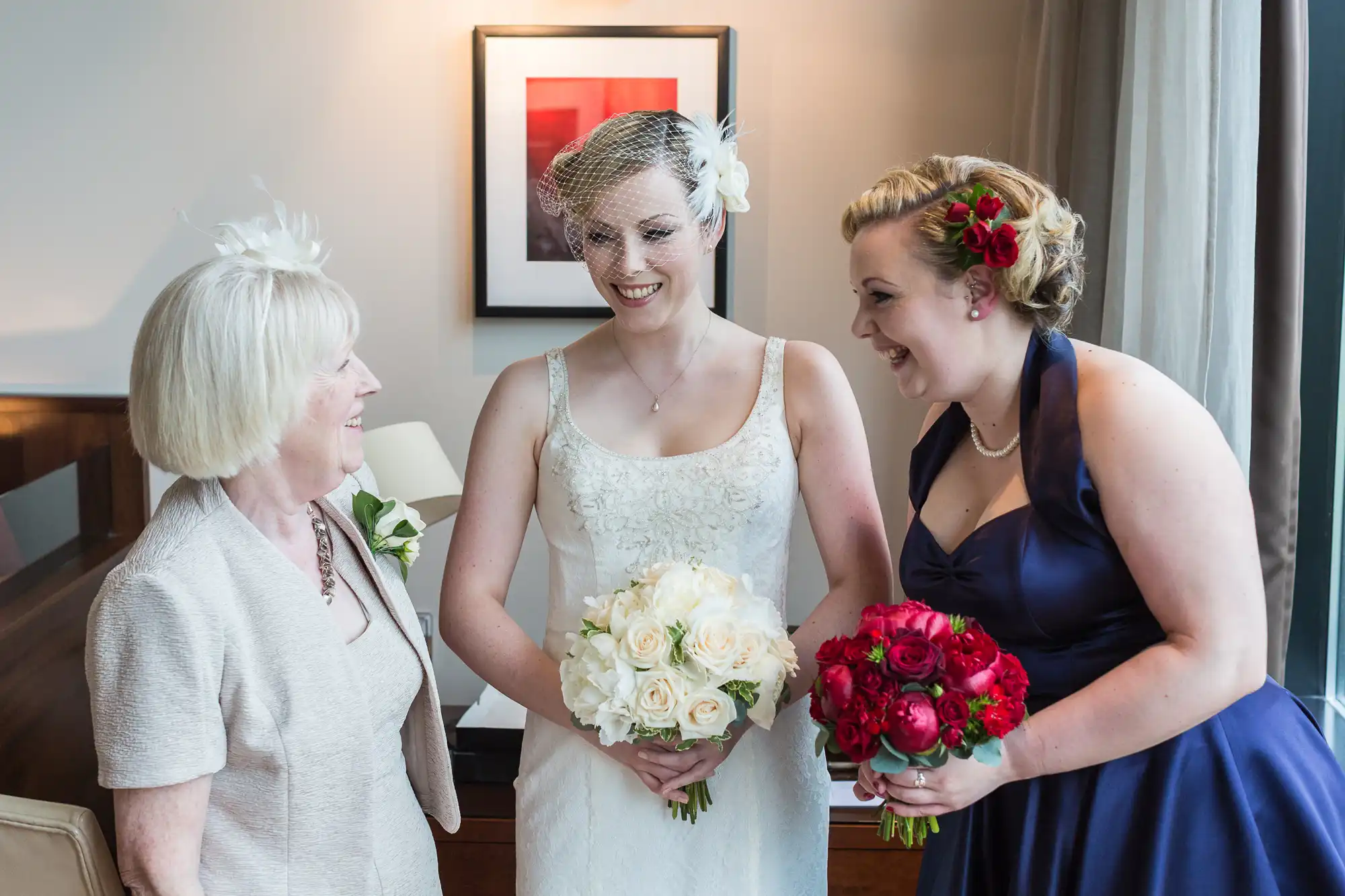 A bride holding a bouquet of white flowers smiles at her bridesmaid with red flowers in her hair, as an older woman in a white dress looks on indoors.