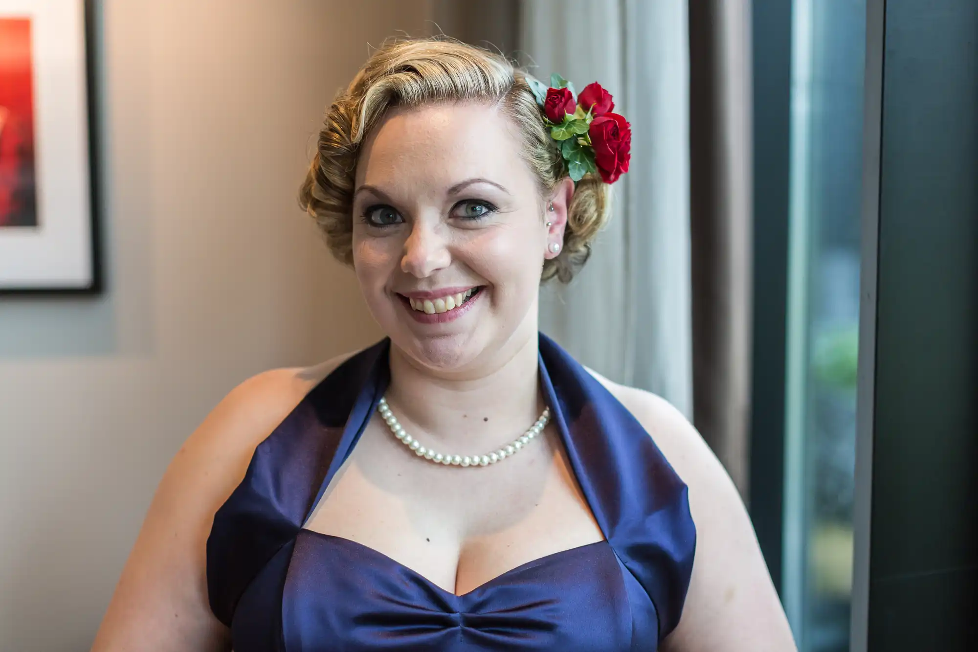 A woman in a blue dress with a pearl necklace, smiling at the camera, her hair styled with red flowers.