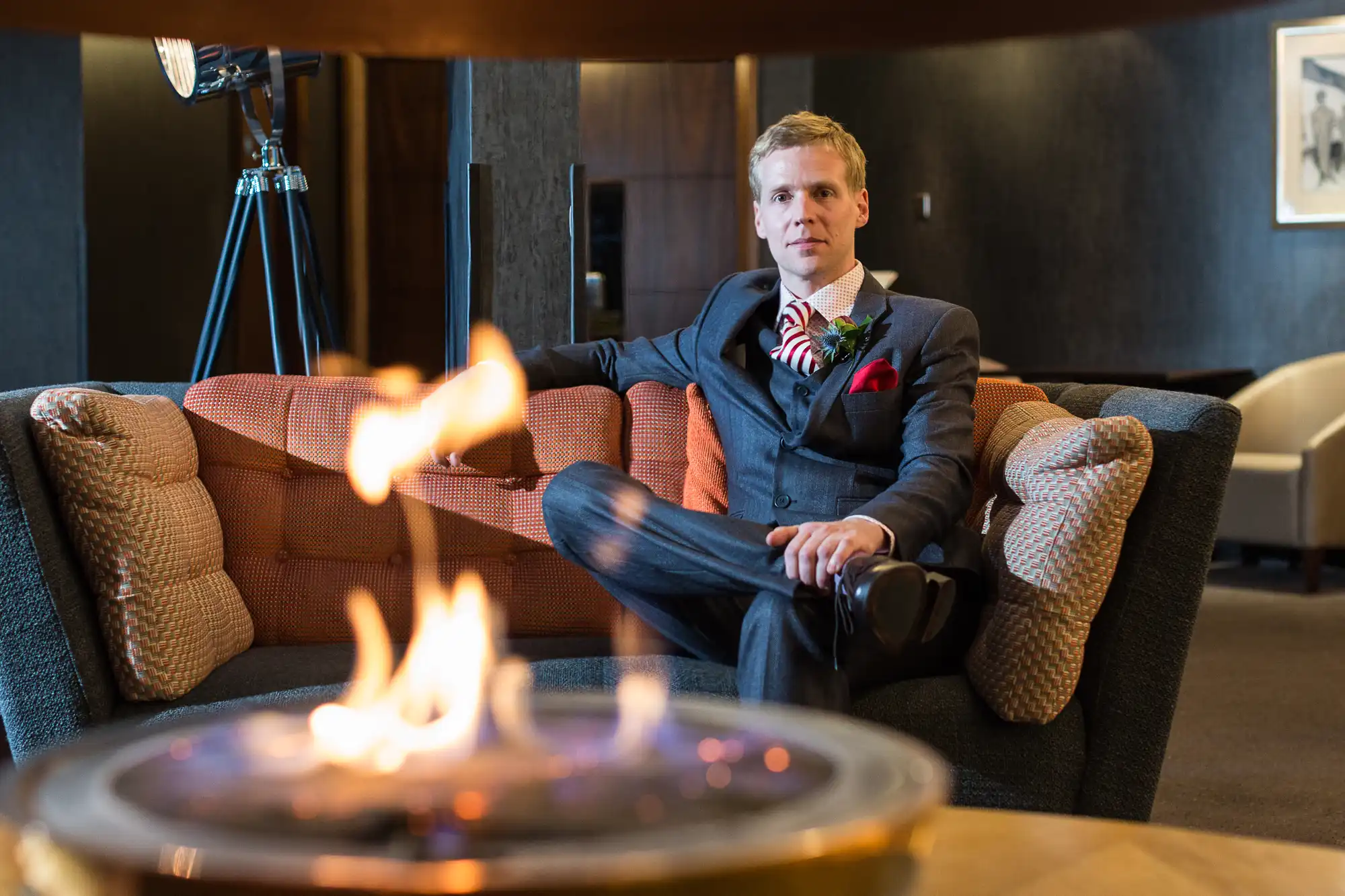 A man in a suit with a boutonniere sits on a couch beside a tabletop fire pit in a stylish, dimly lit lounge.