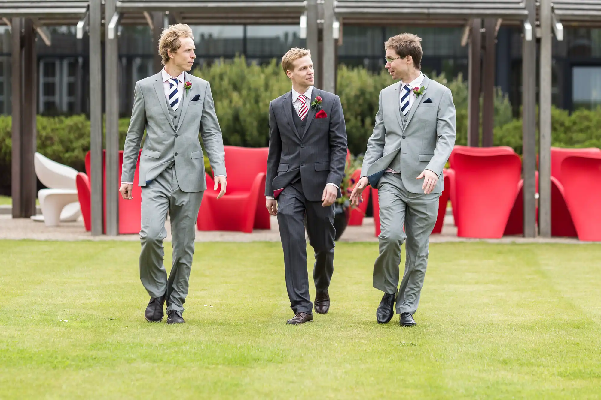 Three men in suits with boutonnieres walk and talk together across a grassy area with modern chairs in the background.