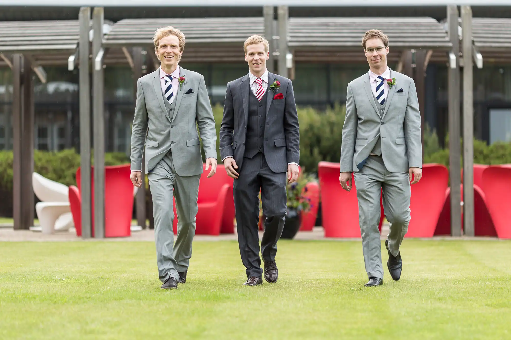 Three men in suits and boutonnieres walking across a lawn, smiling, with modern outdoor furniture in the background.