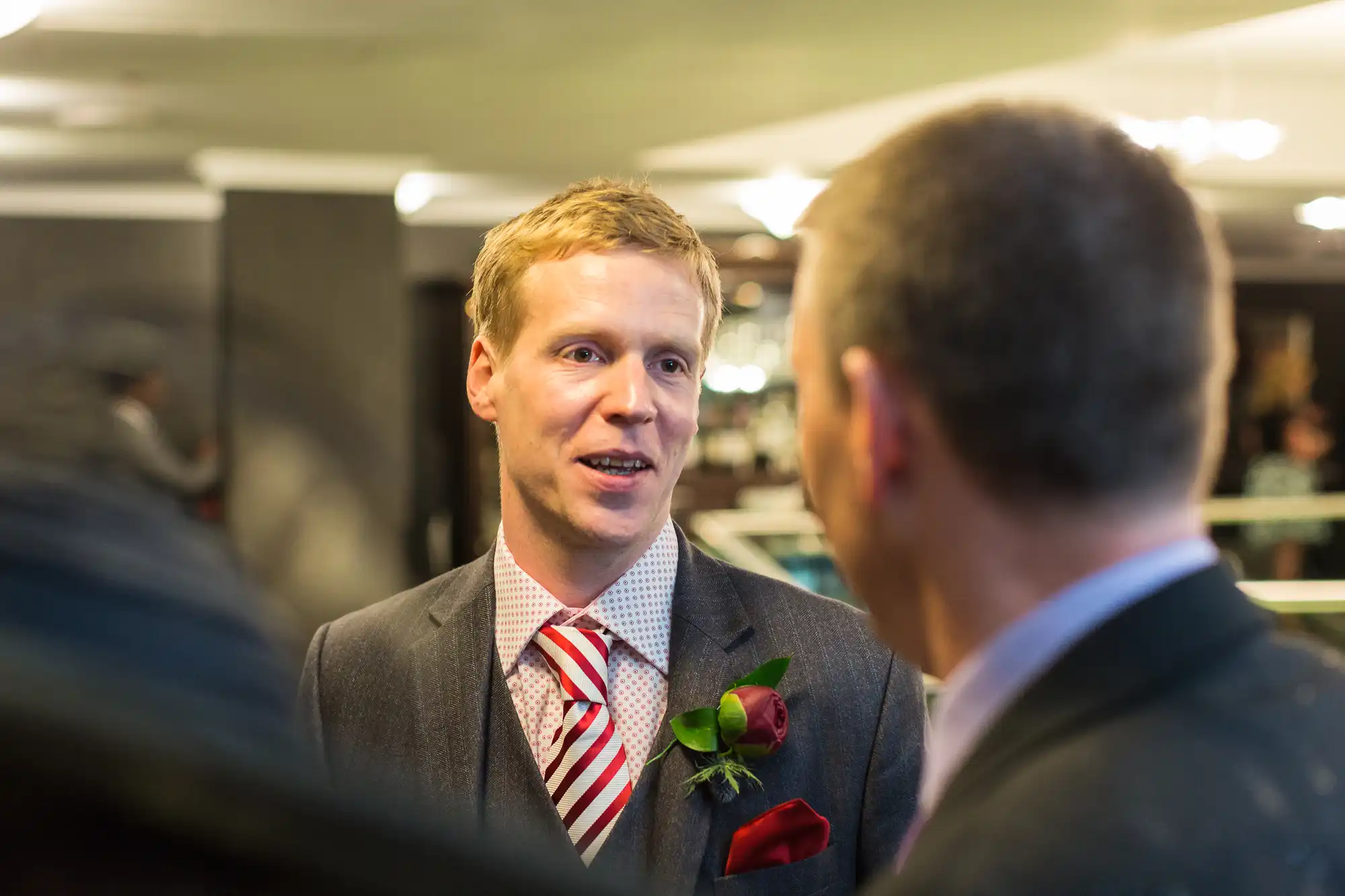 A man in a suit with a striped tie and red boutonniere engaged in conversation at a social event, facing another man whose back is to the camera.