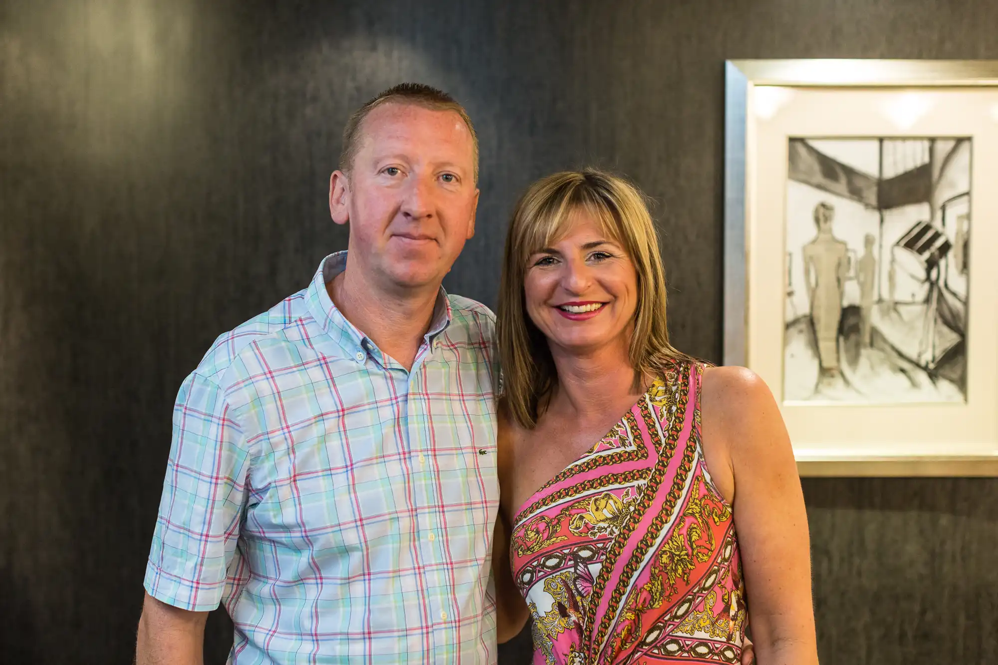 A man and a woman smiling at the camera in a room with a framed sketch on the wall behind them.