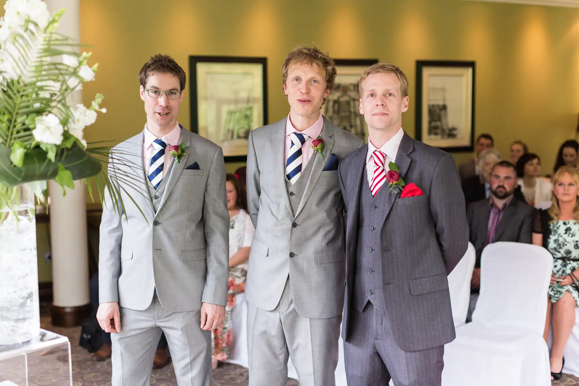Three men in suits at a wedding ceremony, standing in front of seated guests, each wearing a different colored tie and boutonniere.