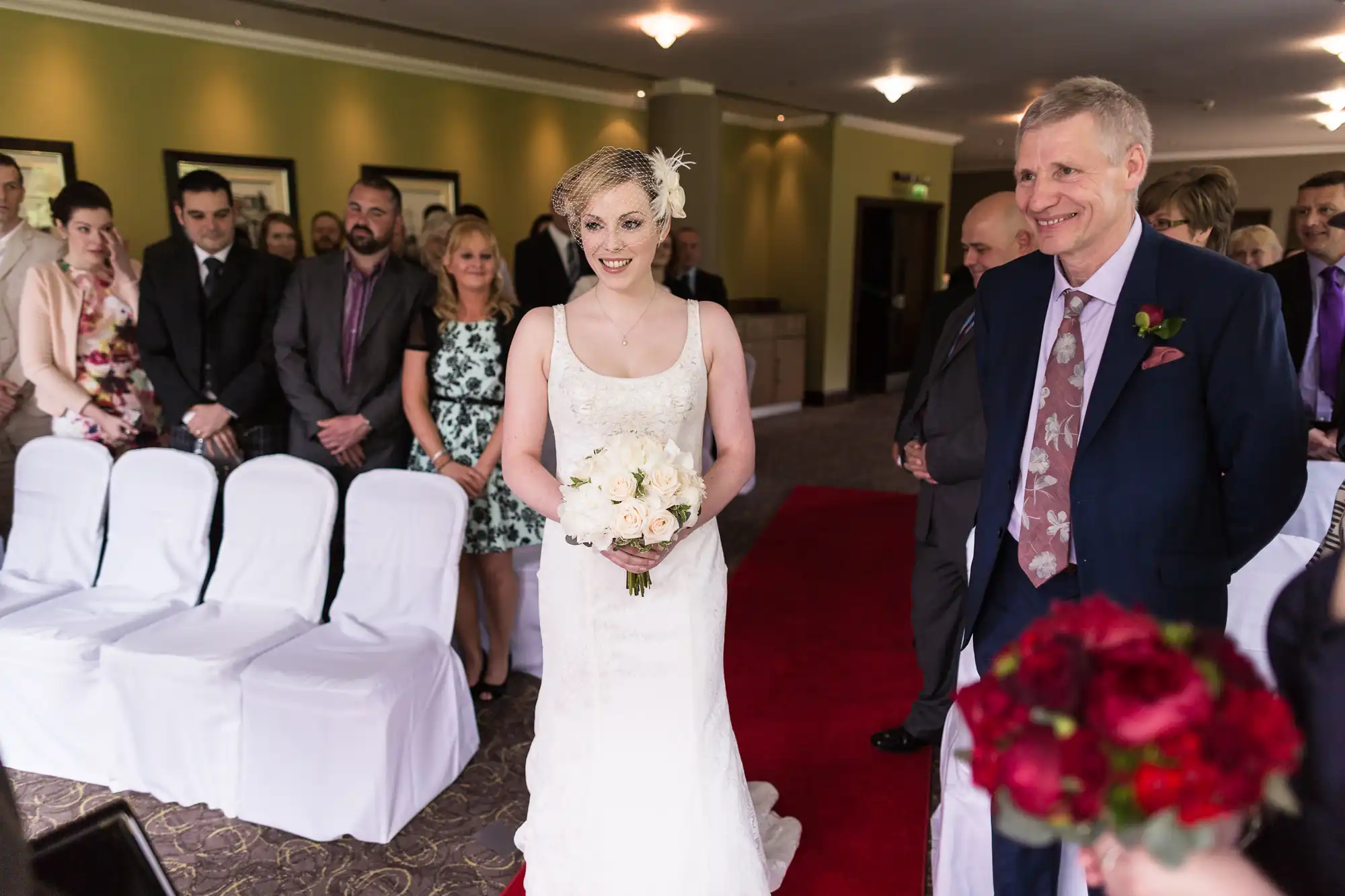 A bride in a white dress and a man in a suit walk down an aisle, smiling, with guests on either side in a wedding hall.