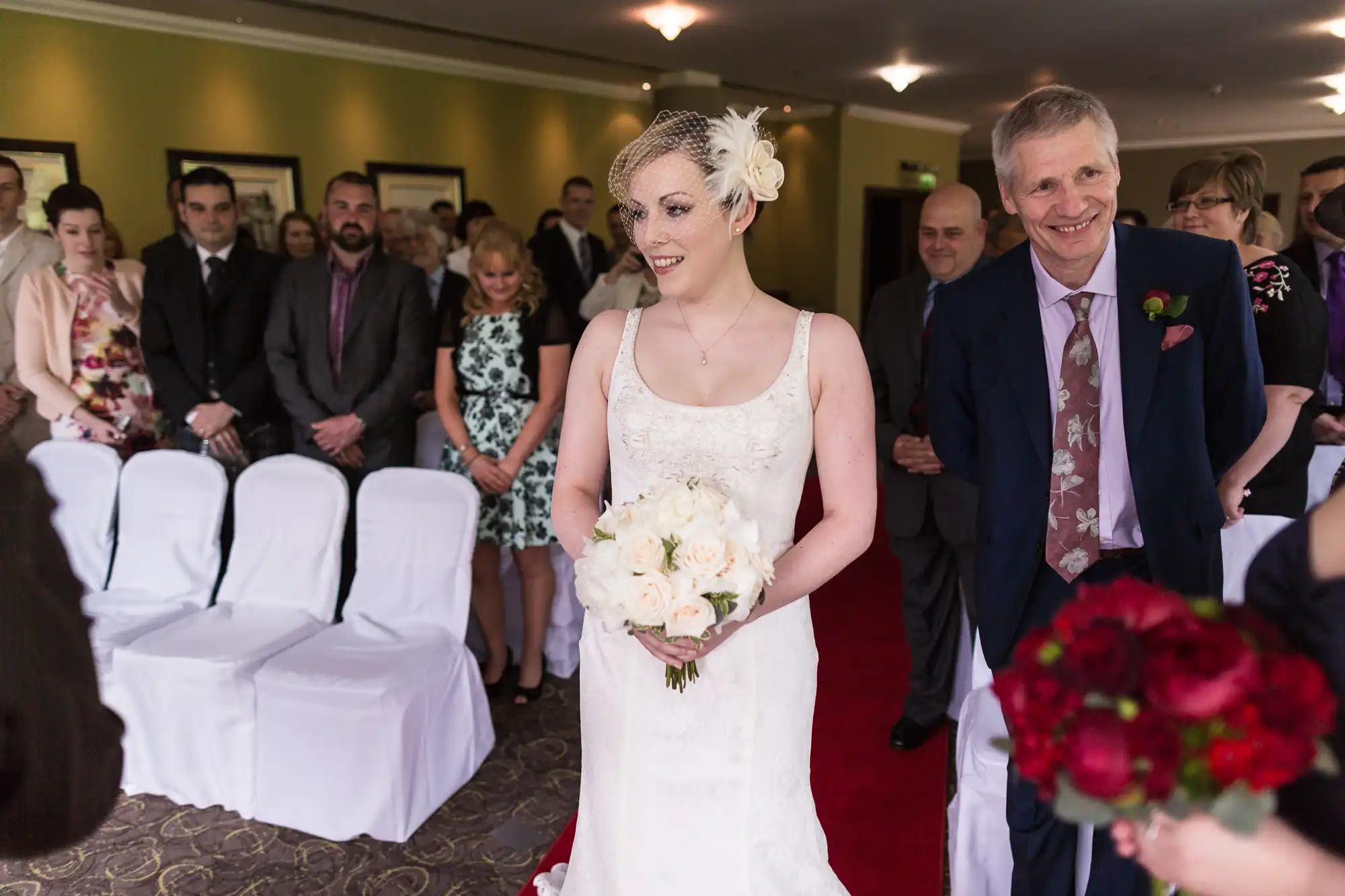 A bride in a white dress with a bouquet walks down the aisle, smiling, accompanied by an older man in a suit, with guests watching.
