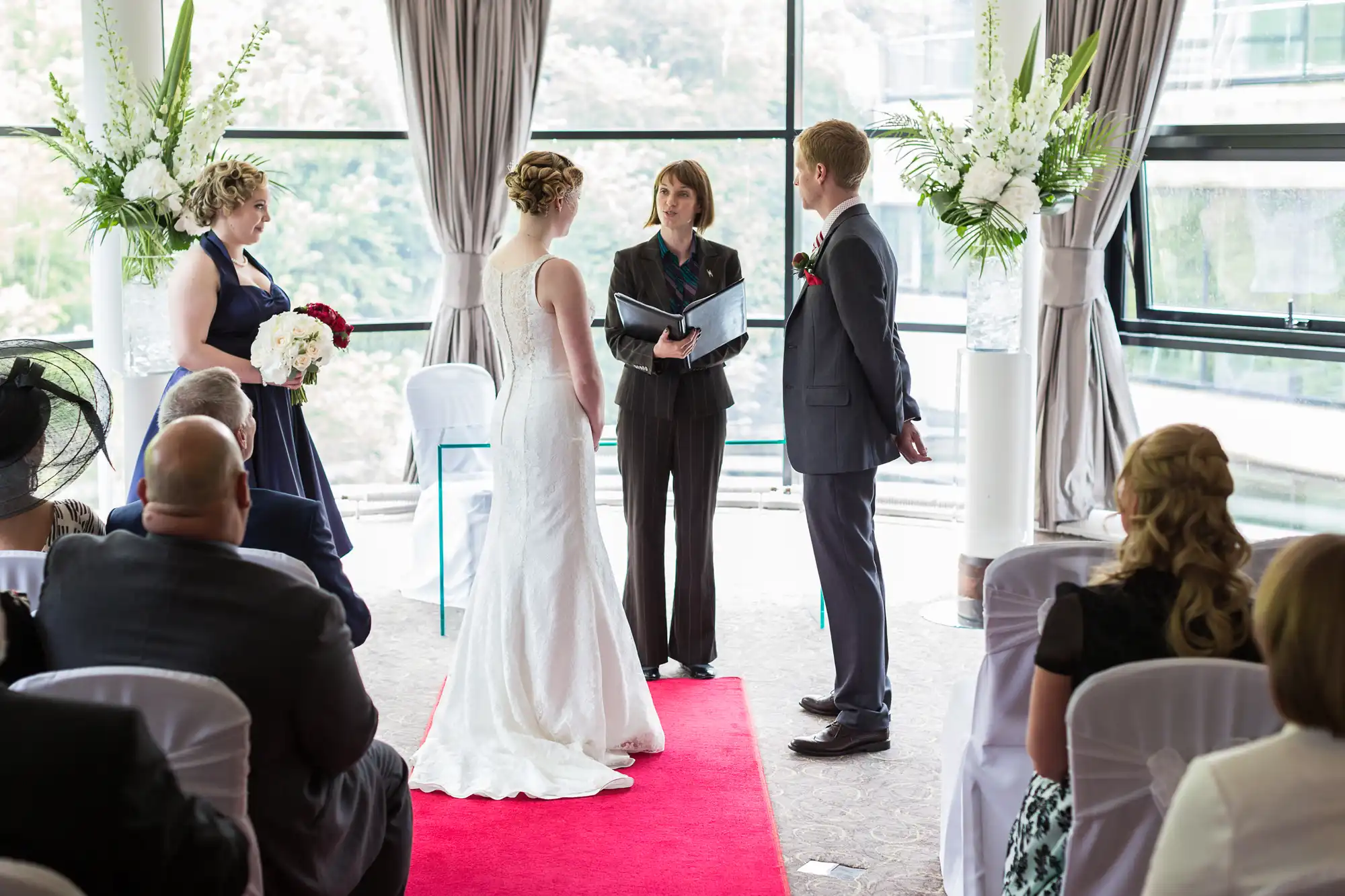 A wedding ceremony indoors with the couple standing at the altar facing the officiant, guests seated around, and large windows in the background.
