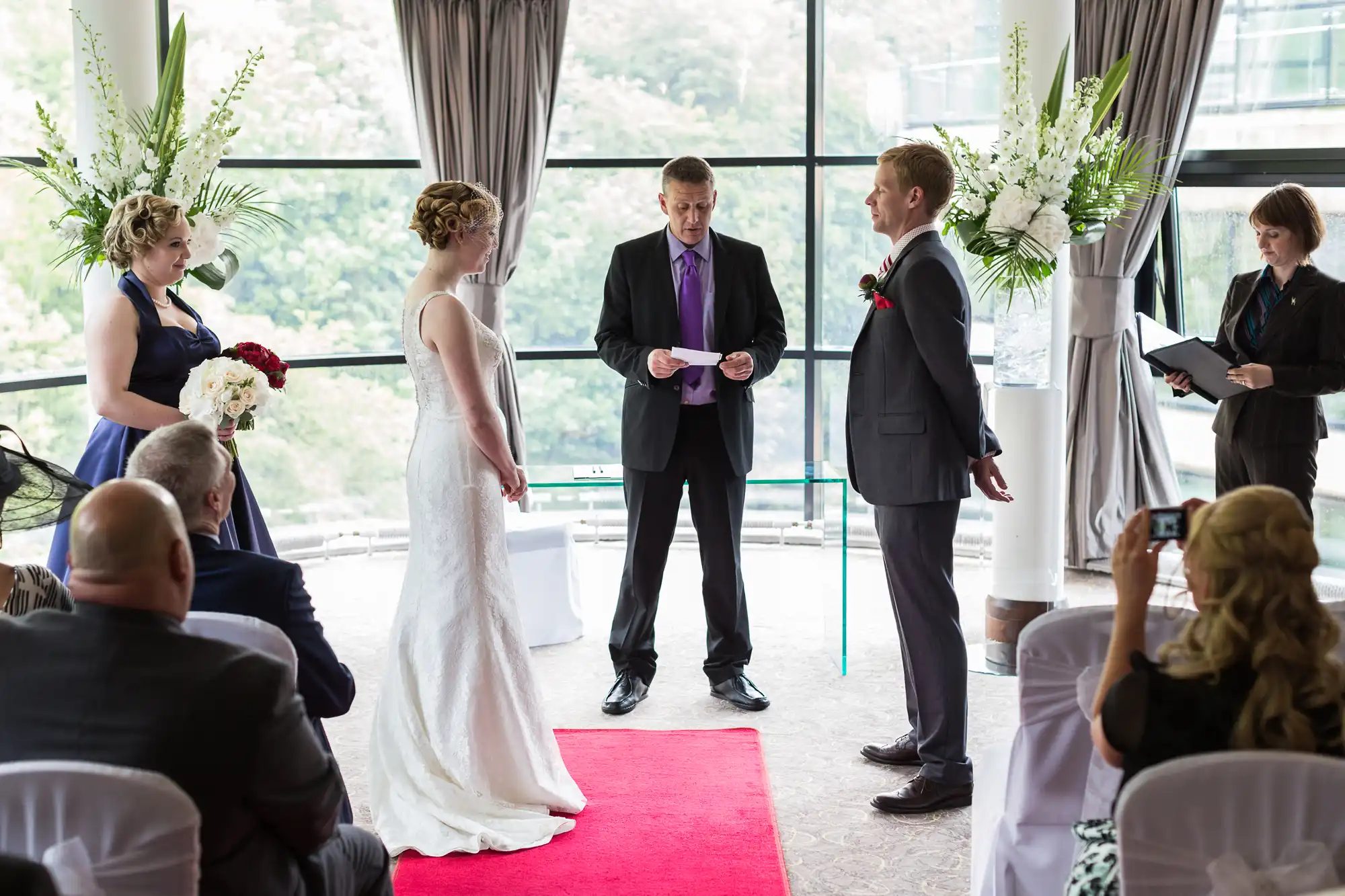 A wedding ceremony in a room with large windows, featuring a bride and a groom facing each other, an officiant reading, and guests watching.
