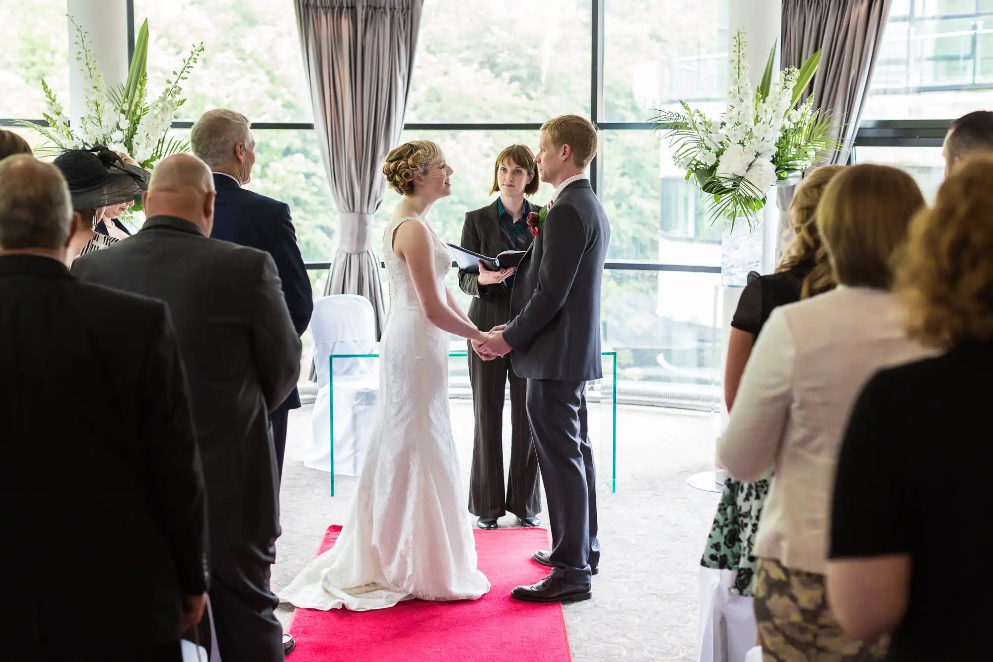 A bride and groom exchange vows at a wedding ceremony indoors, surrounded by guests and decorated with tall floral arrangements.