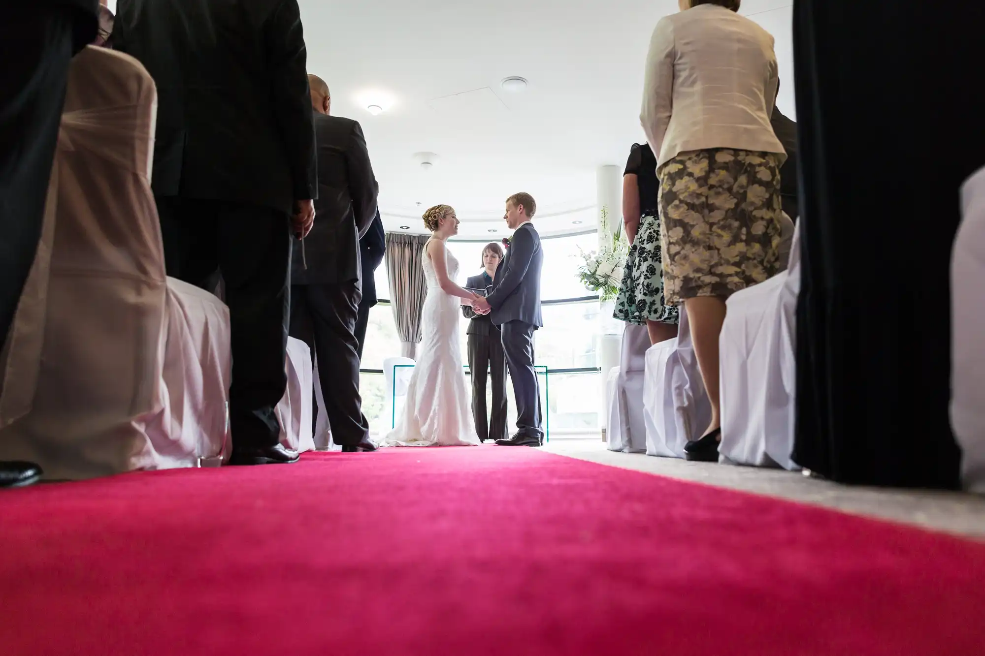 A bride and groom hold hands during a wedding ceremony, viewed from a low angle along a red carpet aisle, surrounded by guests standing.