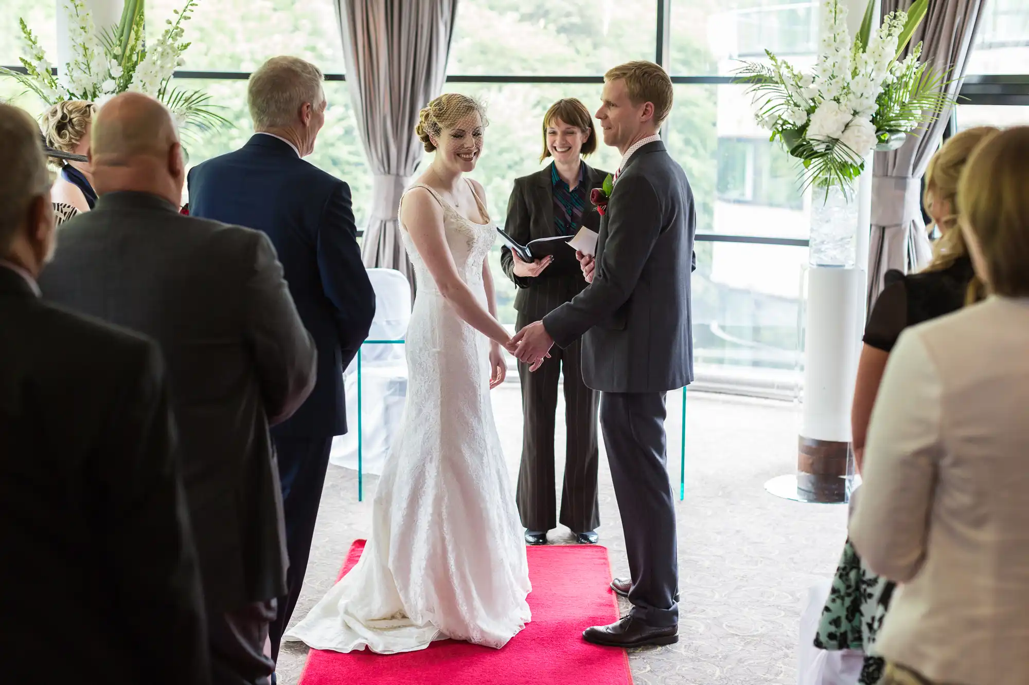 A bride and groom holding hands at their wedding ceremony, standing on a red carpet with an officiant and guests surrounding them.