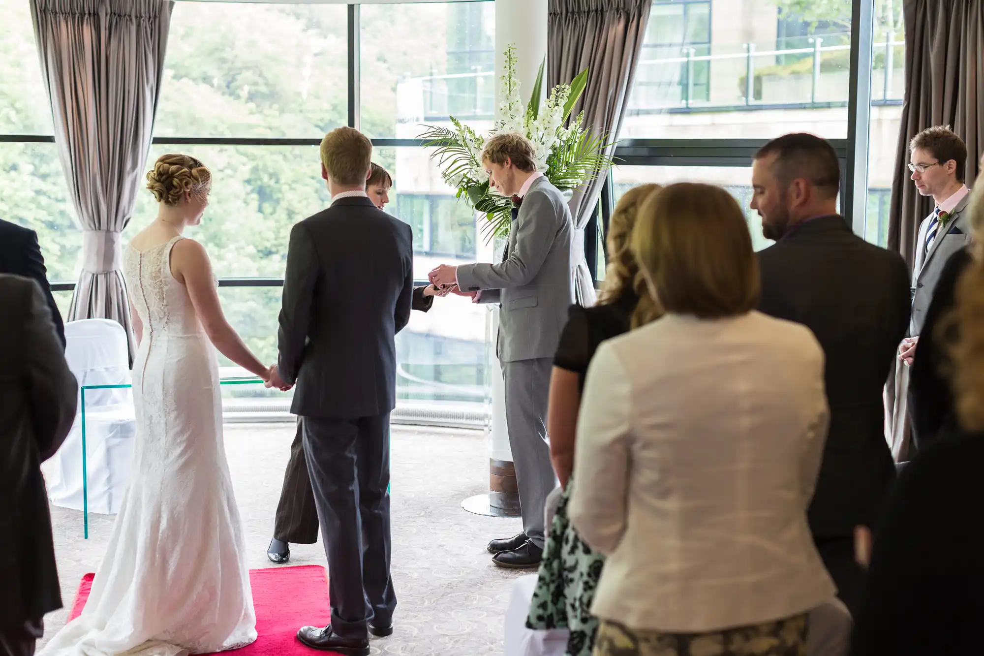 Wedding ceremony in a well-lit room with large windows, where a couple exchanges vows in front of guests.