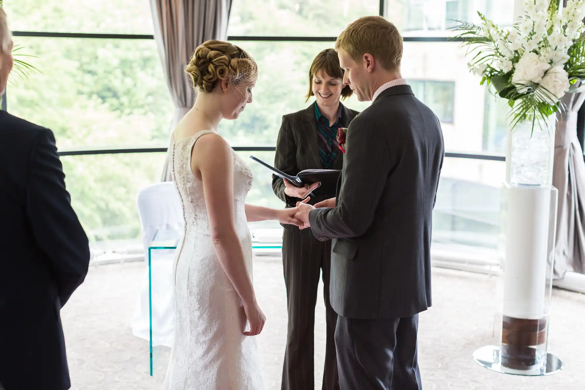 A couple exchanges rings during a wedding ceremony, officiated by a woman, in a room with large windows and a floral arrangement.