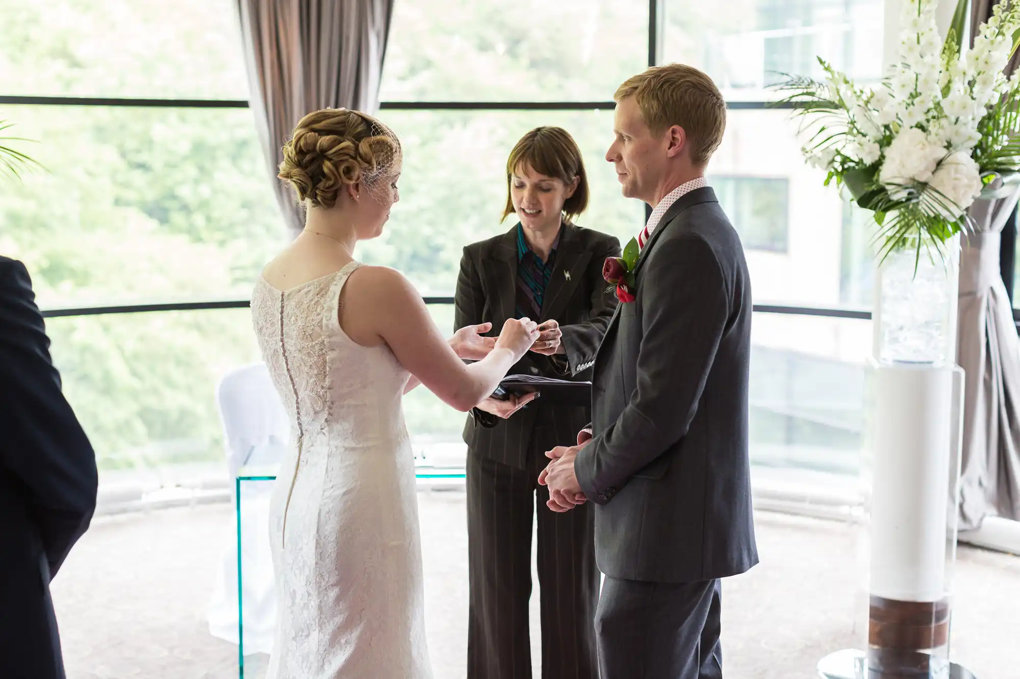 Bride and groom exchanging rings during a wedding ceremony, officiated by a woman, in a room with large windows overlooking greenery.
