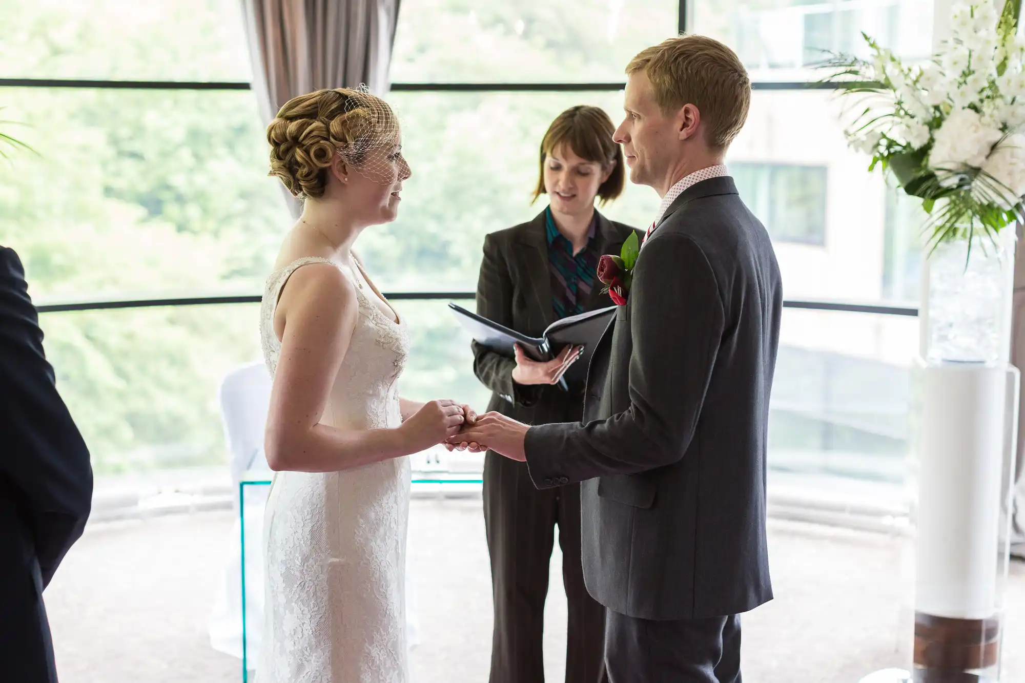 A bride and groom hold hands while exchanging vows, with an officiant holding a book in the background at an indoor wedding venue.