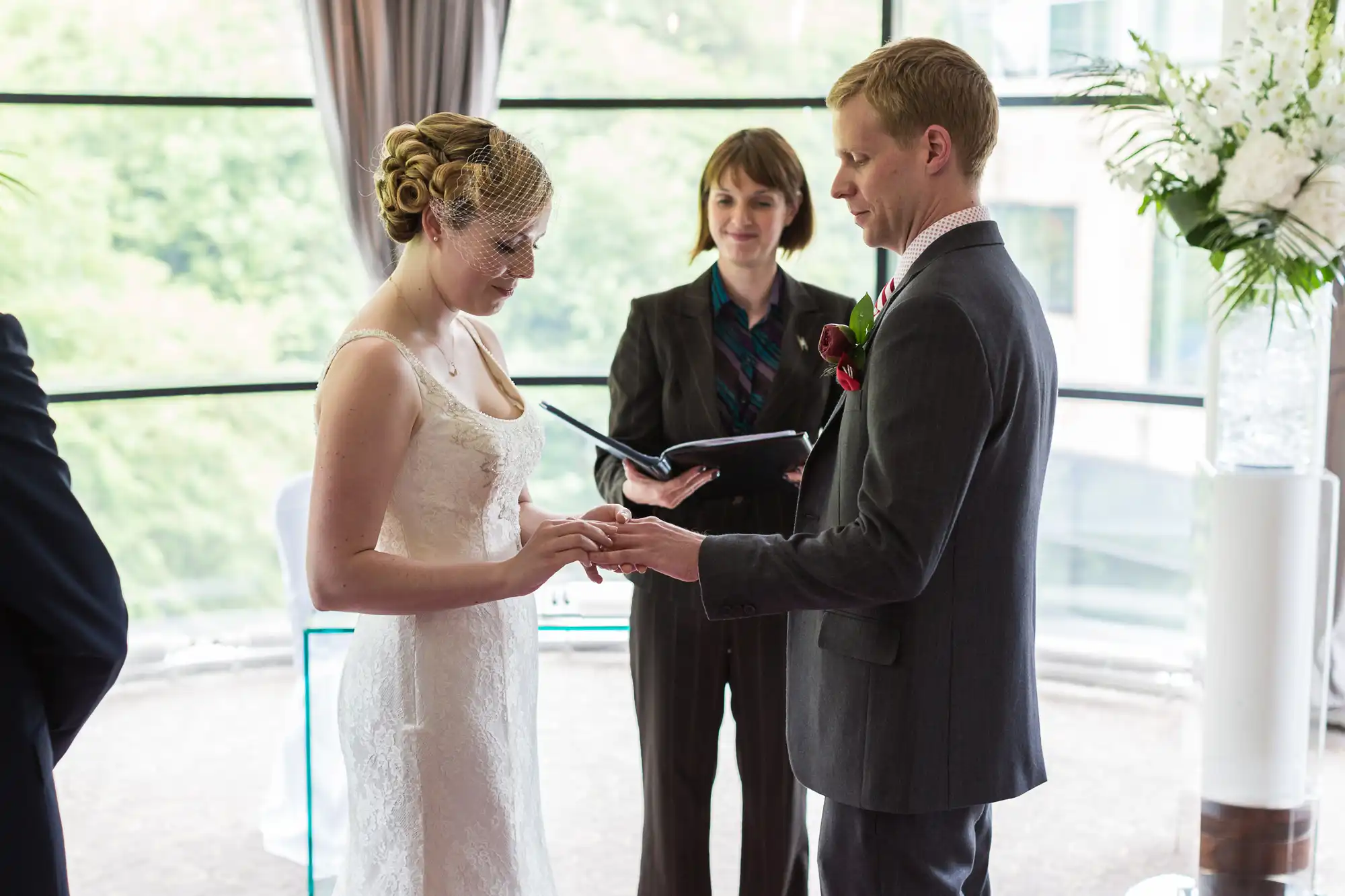 A bride and groom exchange rings during their wedding ceremony, with an officiant watching in the background.