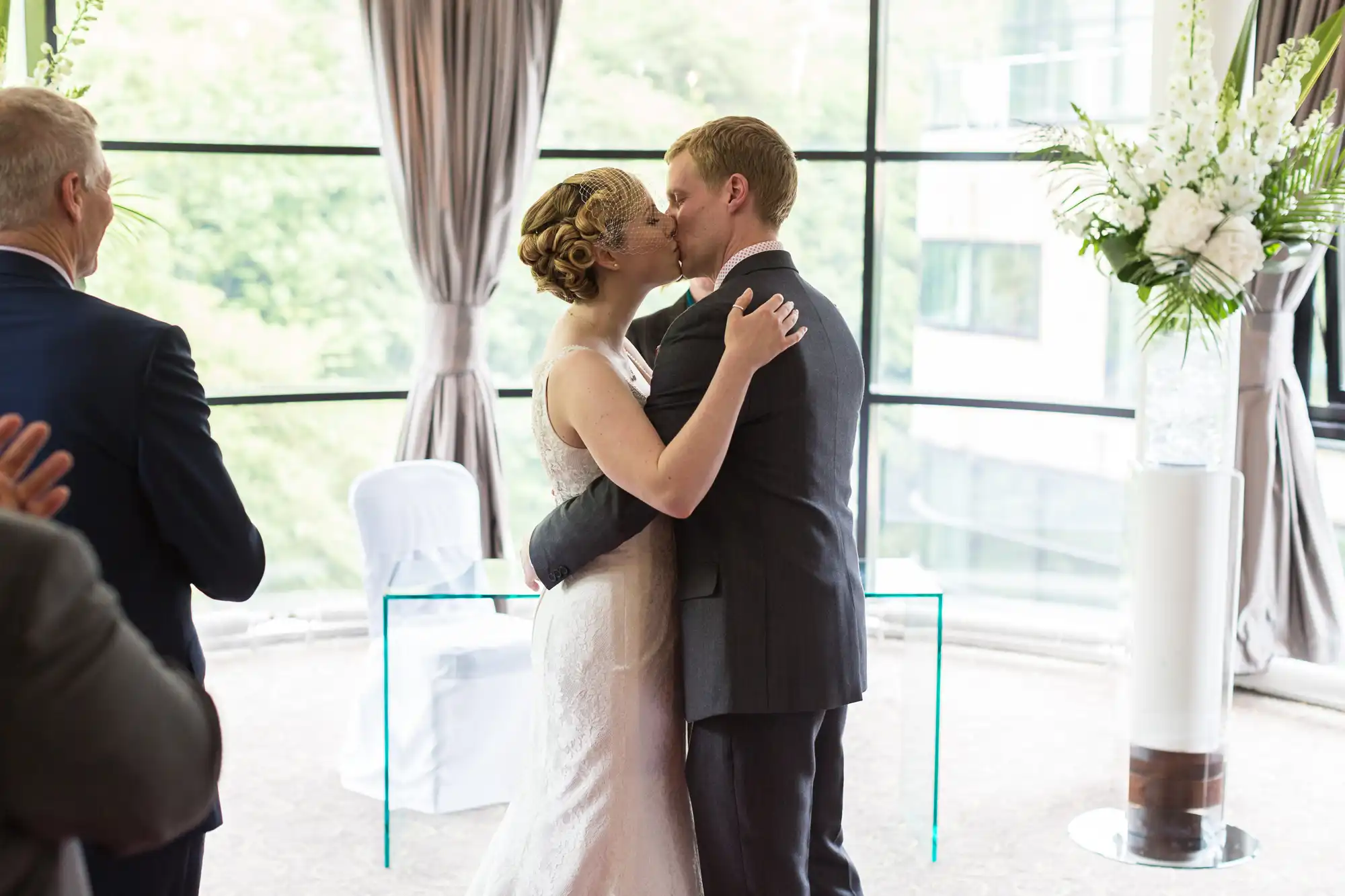 A bride and groom kissing at a wedding reception, surrounded by guests and large windows overlooking trees.