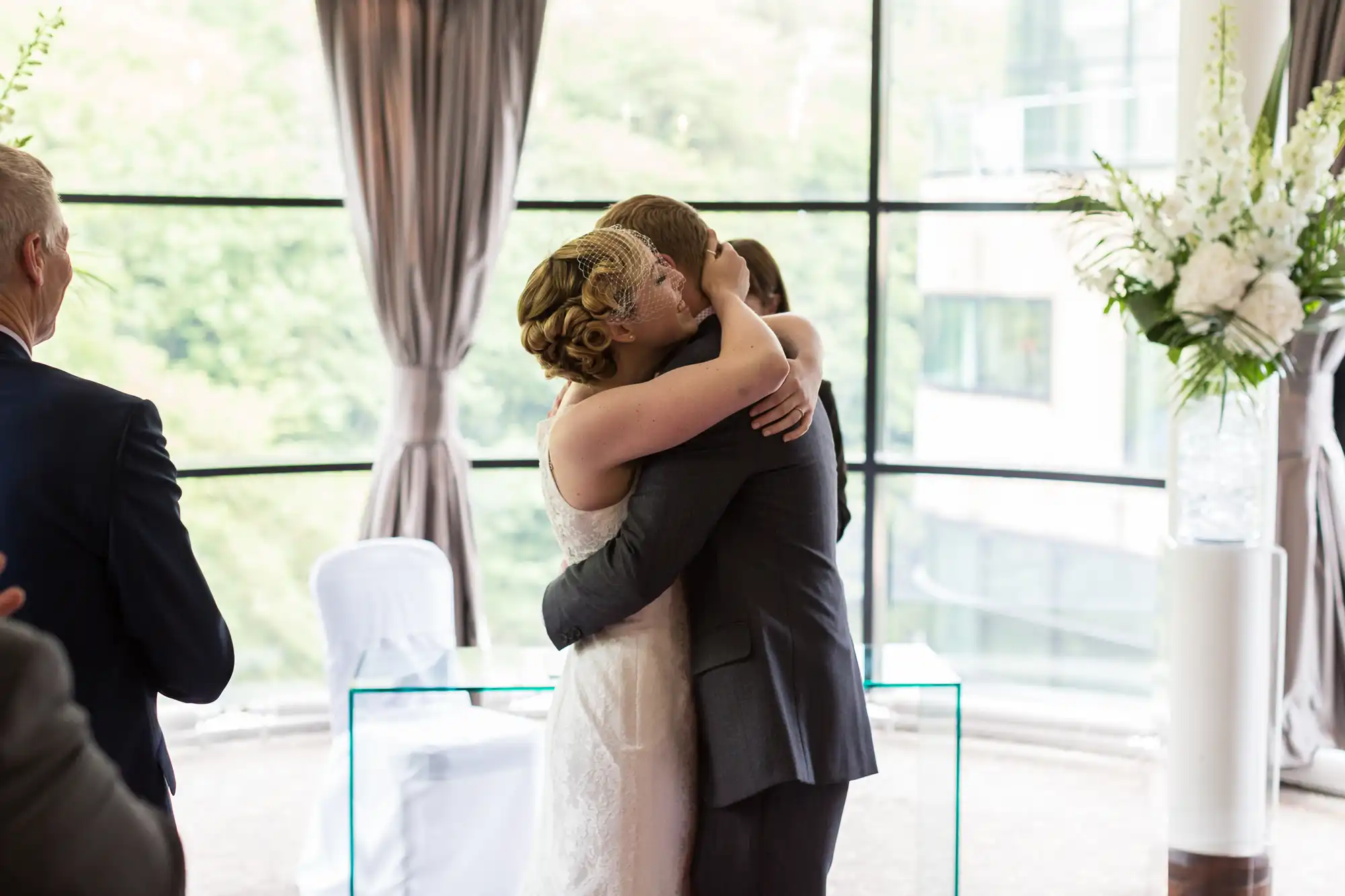 A bride and groom embrace lovingly at their wedding ceremony indoors, with large windows and elegant floral decorations in the background.
