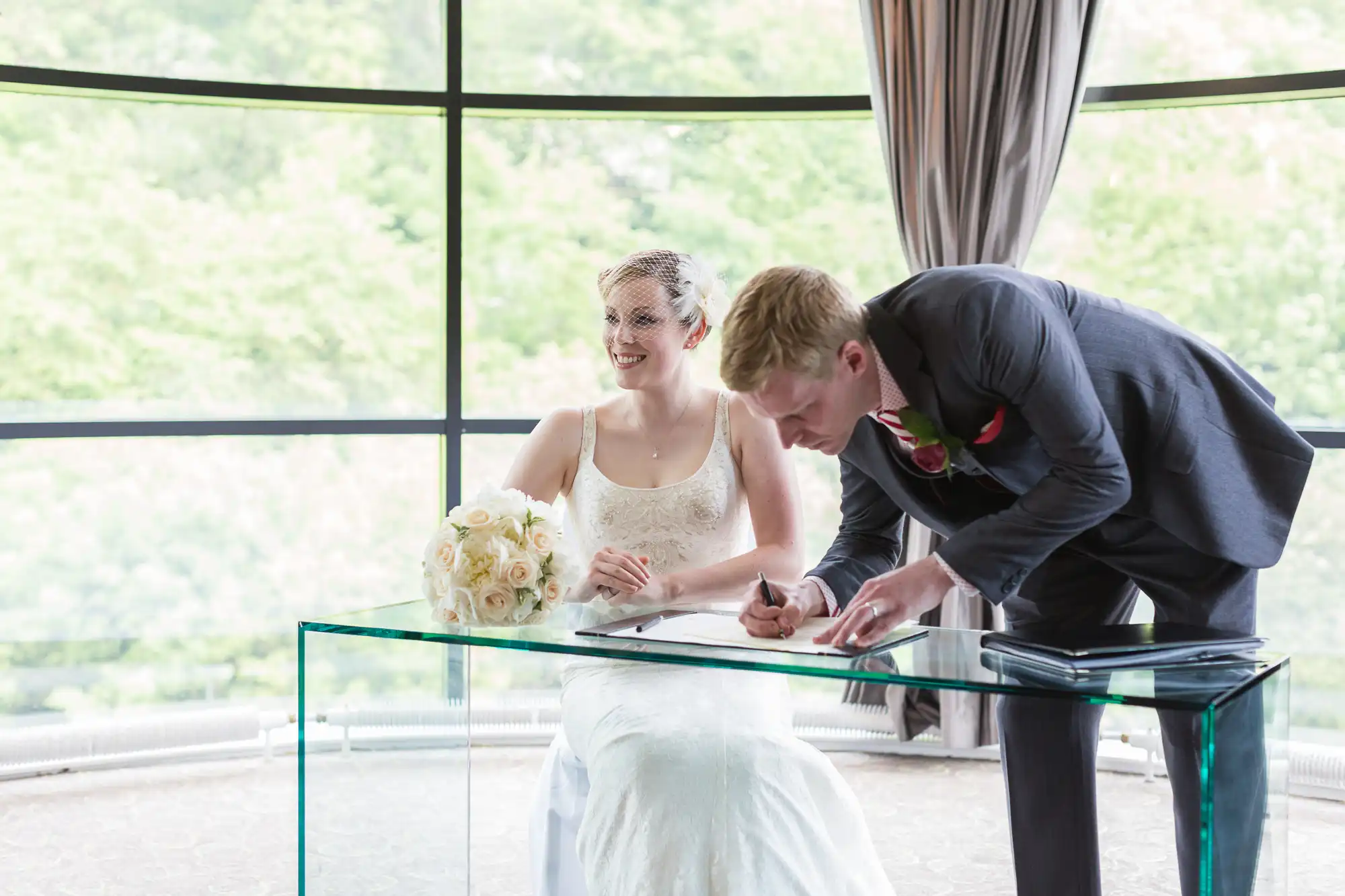 A bride sitting and smiling at the camera as the groom signs a document at a table during a wedding ceremony, with large windows and greenery outside.