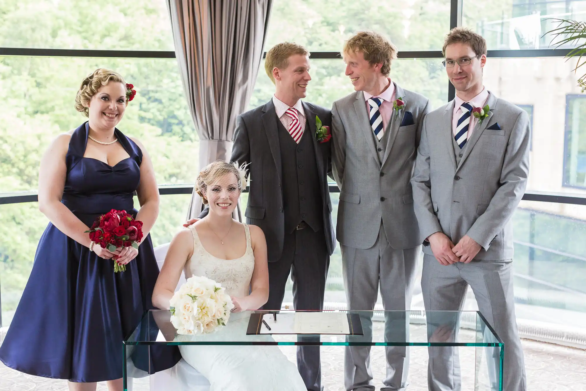 A wedding group indoors with a bride sitting and four others standing, smiling; three men in suits and a woman in a blue dress.