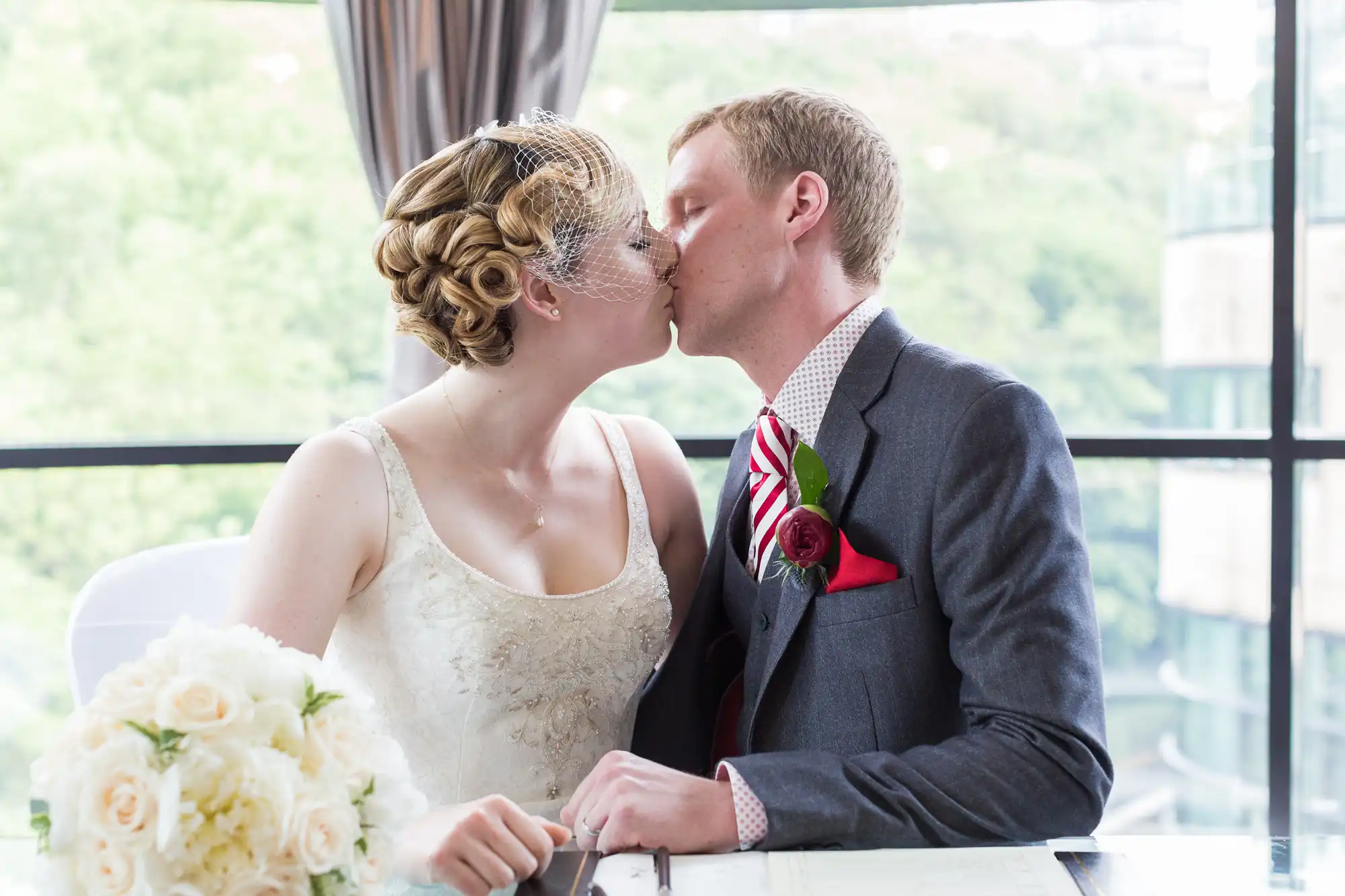 A bride and groom kissing at a table with a bouquet of white roses, in a room with large windows overlooking greenery.