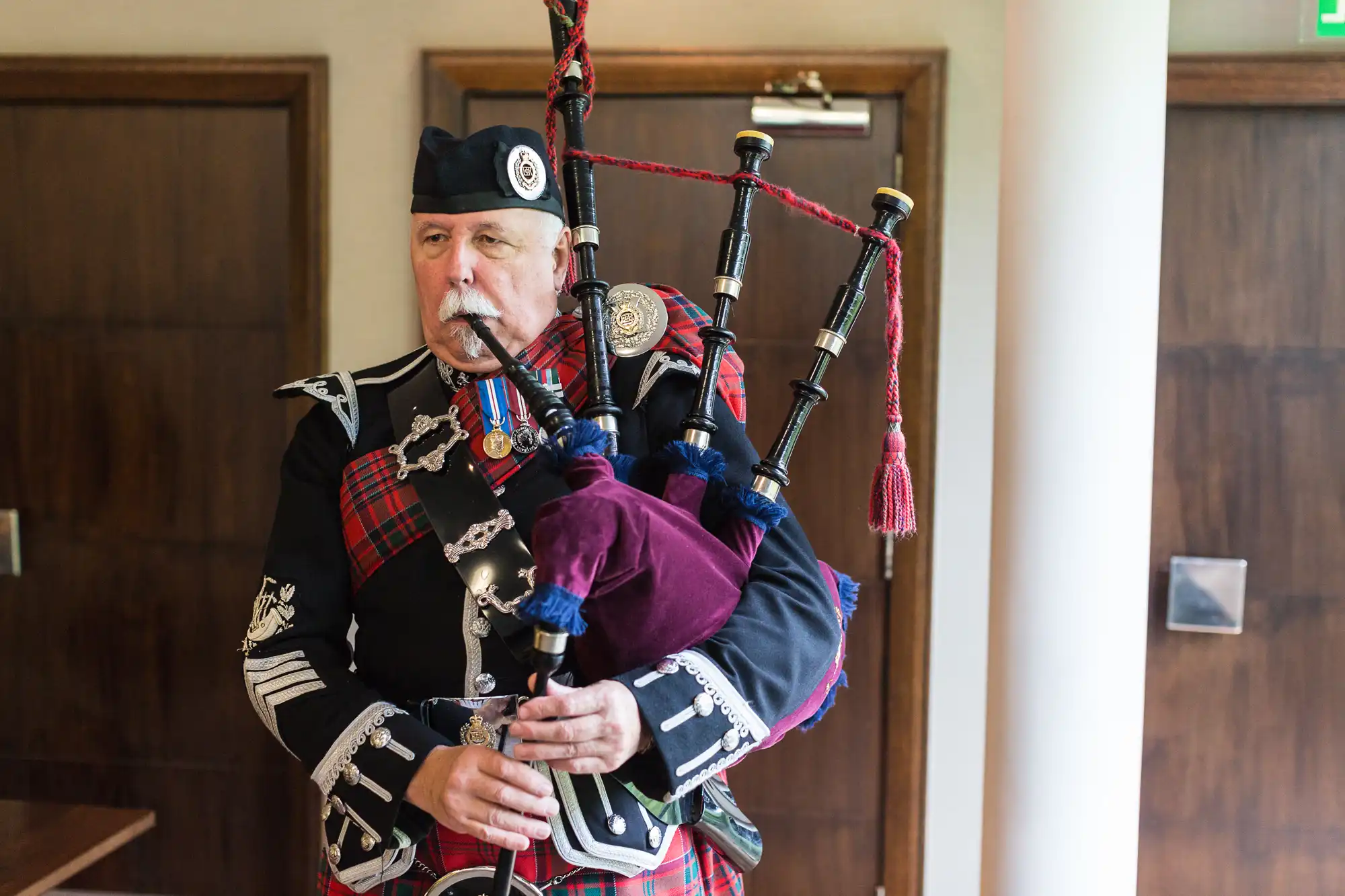 A man in traditional scottish attire, including a kilt and sporran, playing the bagpipes indoors.