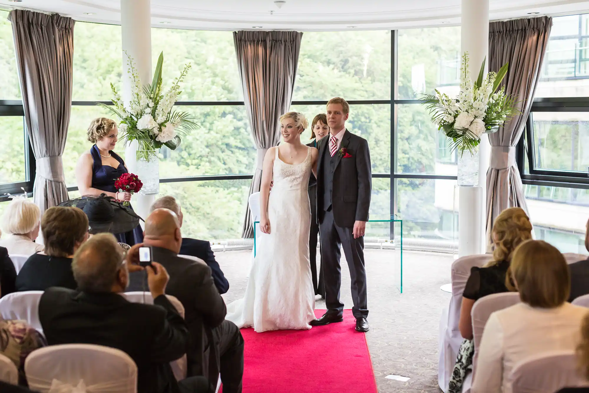 A bride and groom standing at the end of a red carpet in a wedding ceremony, with guests seated around them in a room with large windows and curtains.