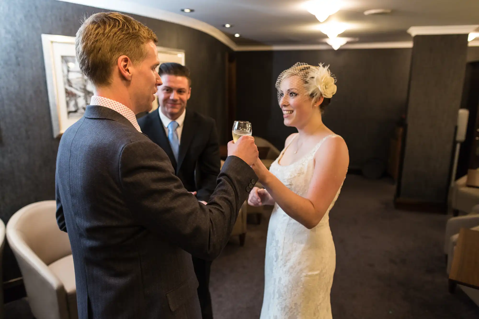 A bride in a white dress with a floral hairpiece and a groom in a suit toast with wine glasses, smiling at each other, with a man observing in the background.