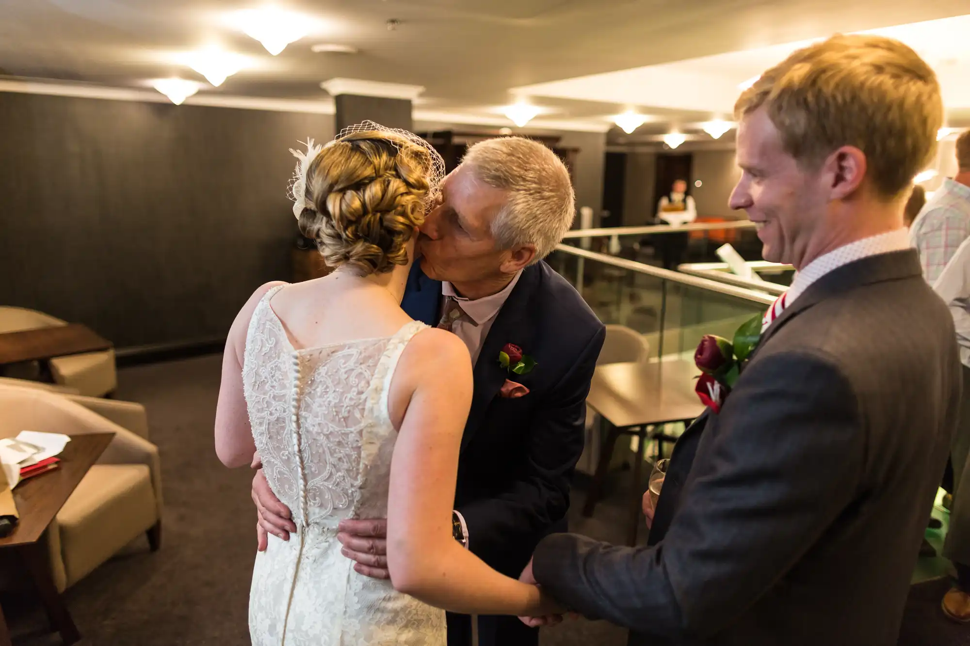A bride in a lace gown receives a kiss on the cheek from an older man while a smiling man watches nearby at an indoor venue.