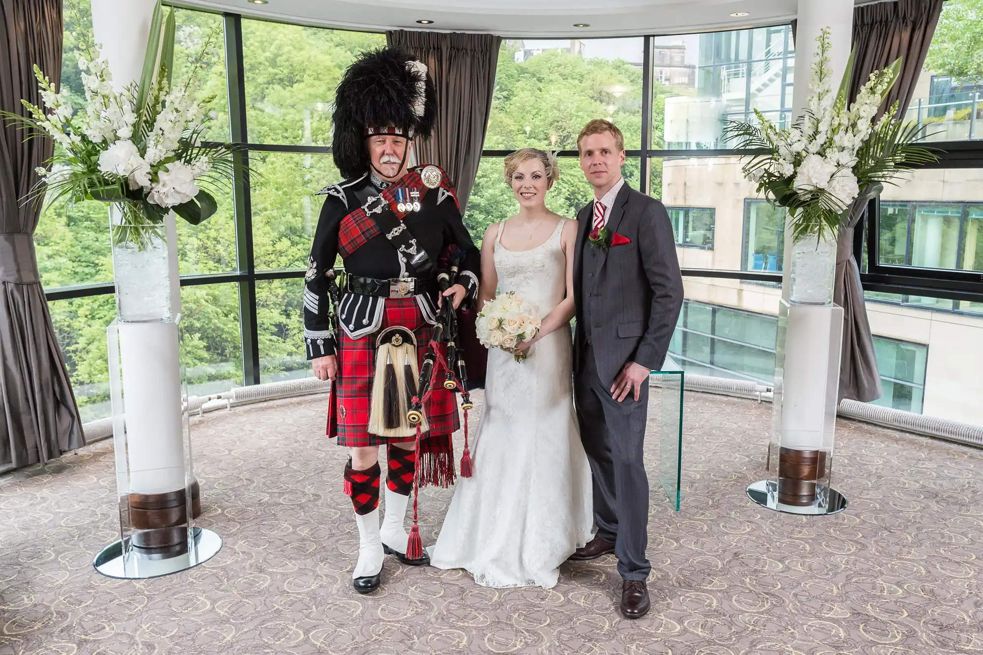A bride and groom posing with a bagpiper in traditional scottish attire at an indoor wedding venue surrounded by windows and floral decorations.