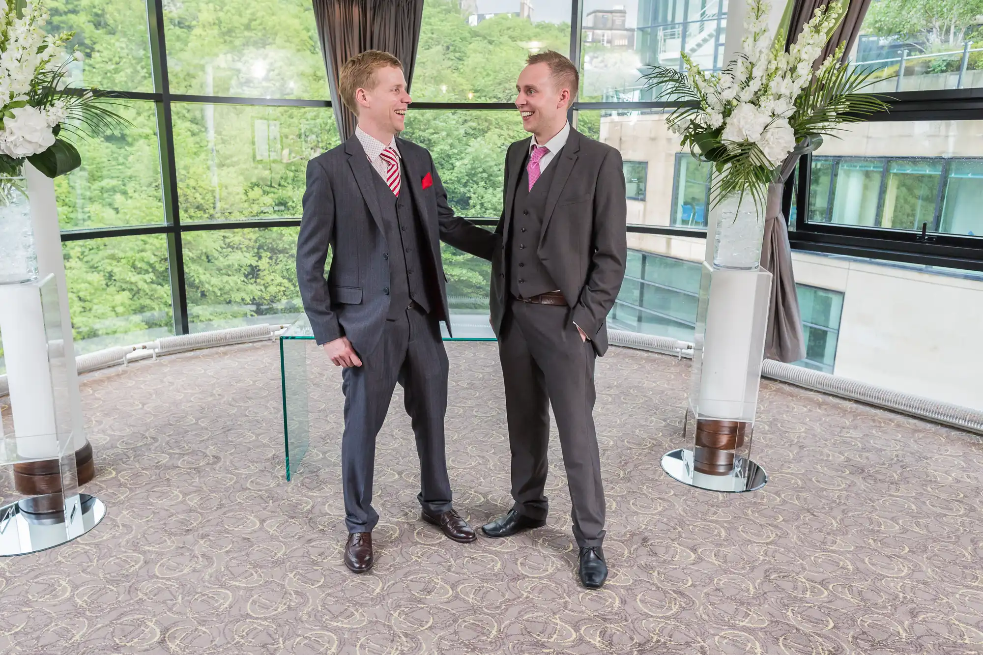Two men smiling and holding hands at a wedding ceremony indoors, with large windows and greenery outside.