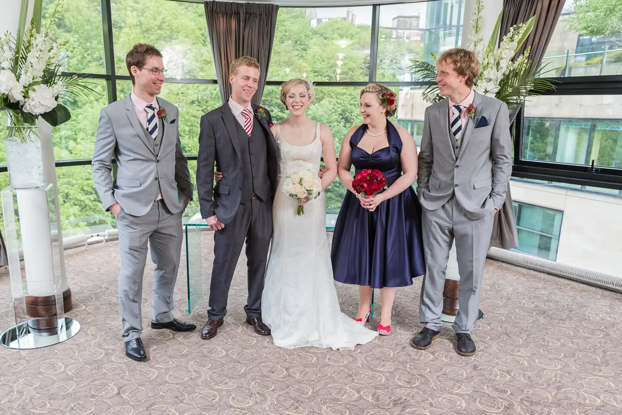 A bride and groom posing happily with three friends in formal attire at an indoor wedding venue with large windows and greenery outside.