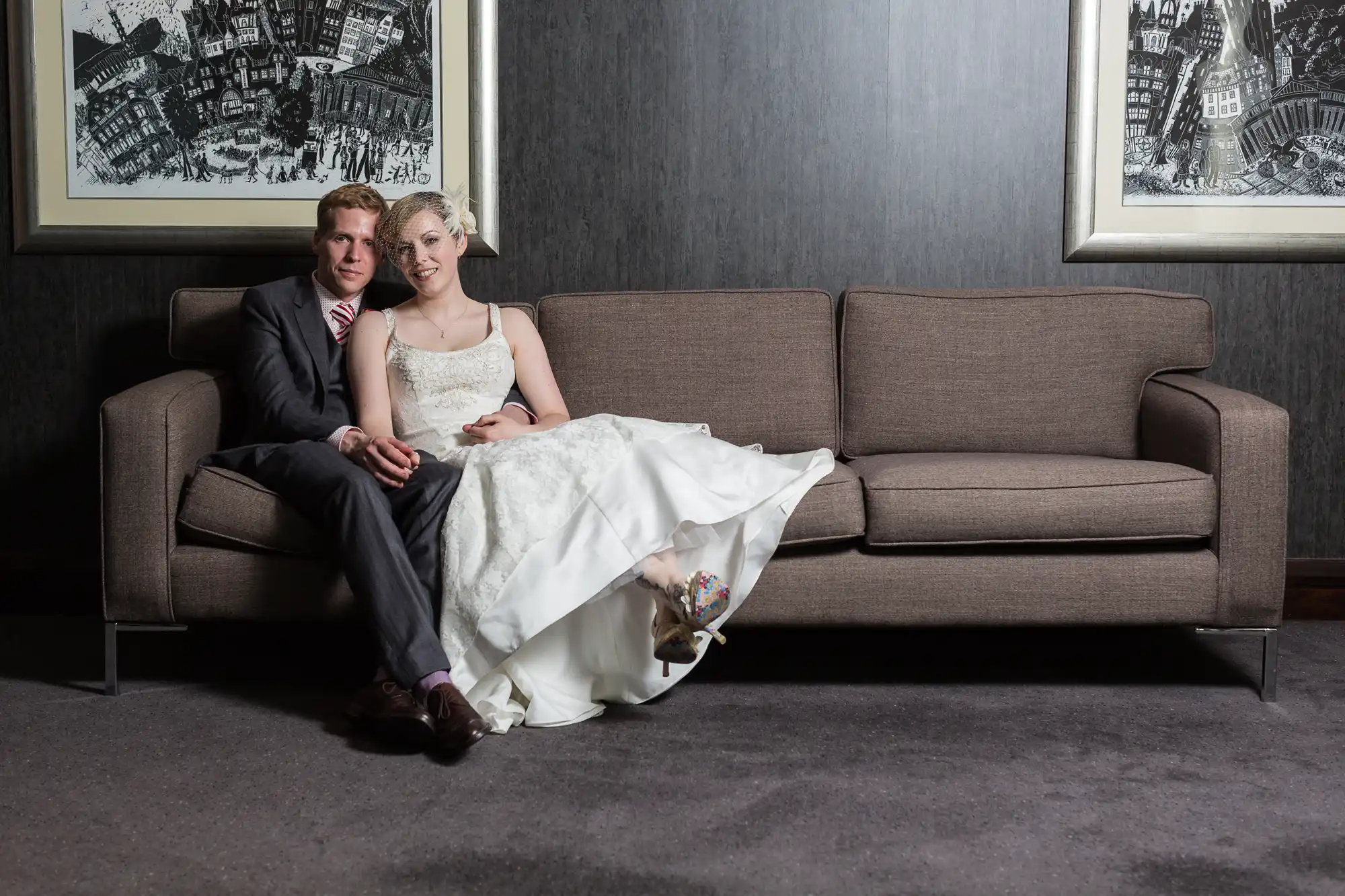 A bride and groom sitting closely on a brown sofa, smiling subtly, in a room with gray walls and framed artwork.