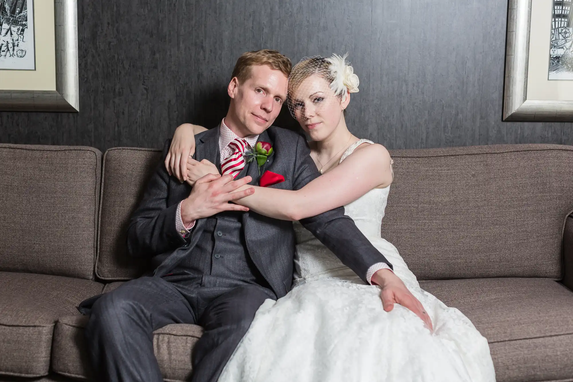 A newlywed couple embracing on a sofa, the groom in a dark suit and the bride in a white gown, both looking directly at the camera.